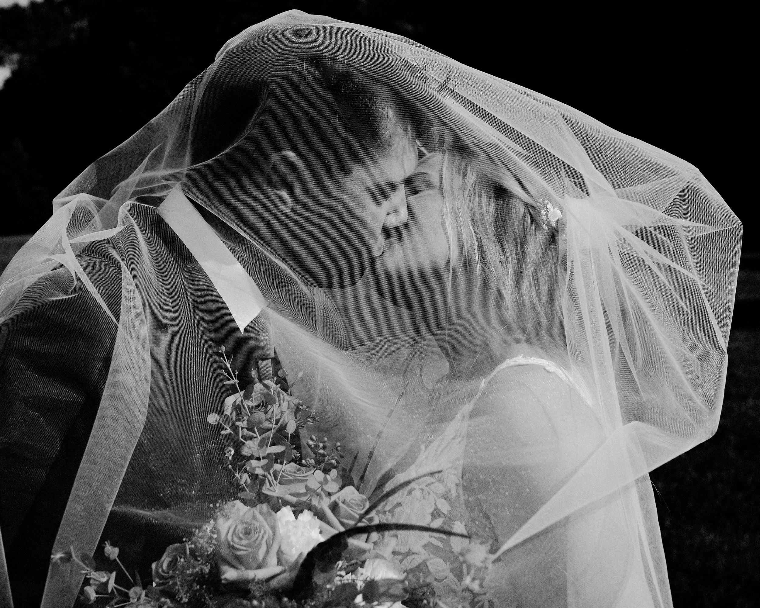 Black and white photo of a bride and groom embracing under a veil, with the bride holding a bouquet of flowers.