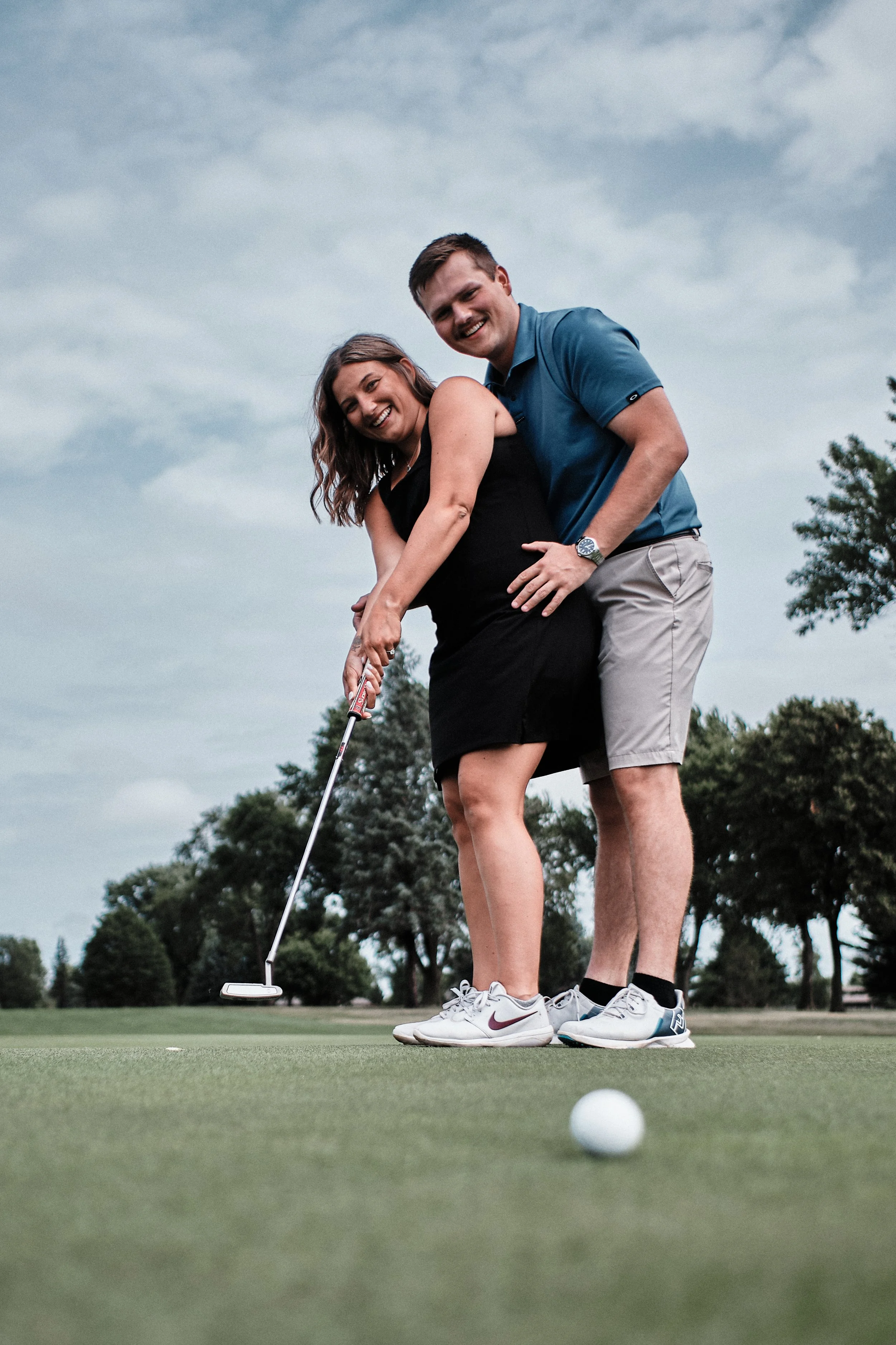 A couple playing golf on a green field, preparing to putt the ball, with trees and cloudy sky in the background.