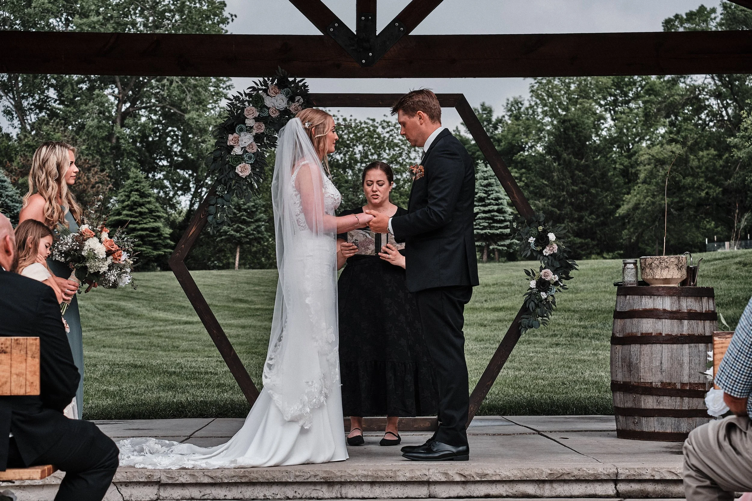 A wedding ceremony is taking place outdoors on a grassy area with trees in the background. The bride and groom are holding hands and looking at each other beneath a wooden arch decorated with flowers. The officiant is standing between them, reading f