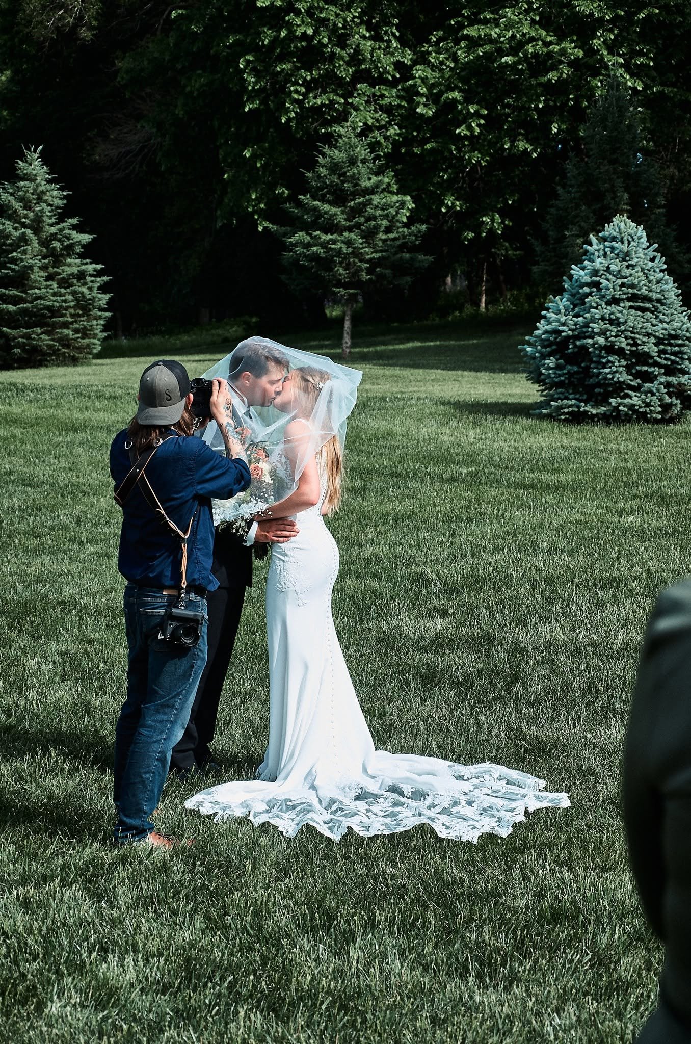 A bride and groom sharing a kiss outdoors while a photographer captures the moment. The bride is wearing a white wedding dress and veil, holding a bouquet, with a long train on the grass. The scene is set in a lush green park with trees in the background.