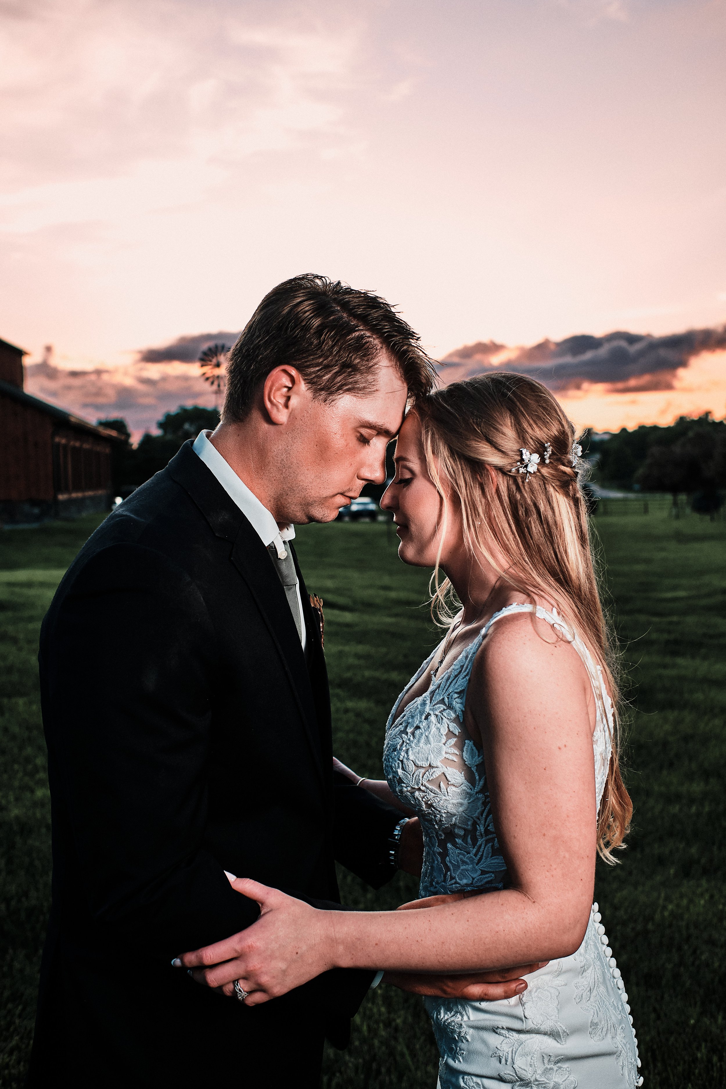 A bride and groom with foreheads touching during sunset at a farm, holding hands, dressed in wedding attire, with a barn and a windmill in the background.