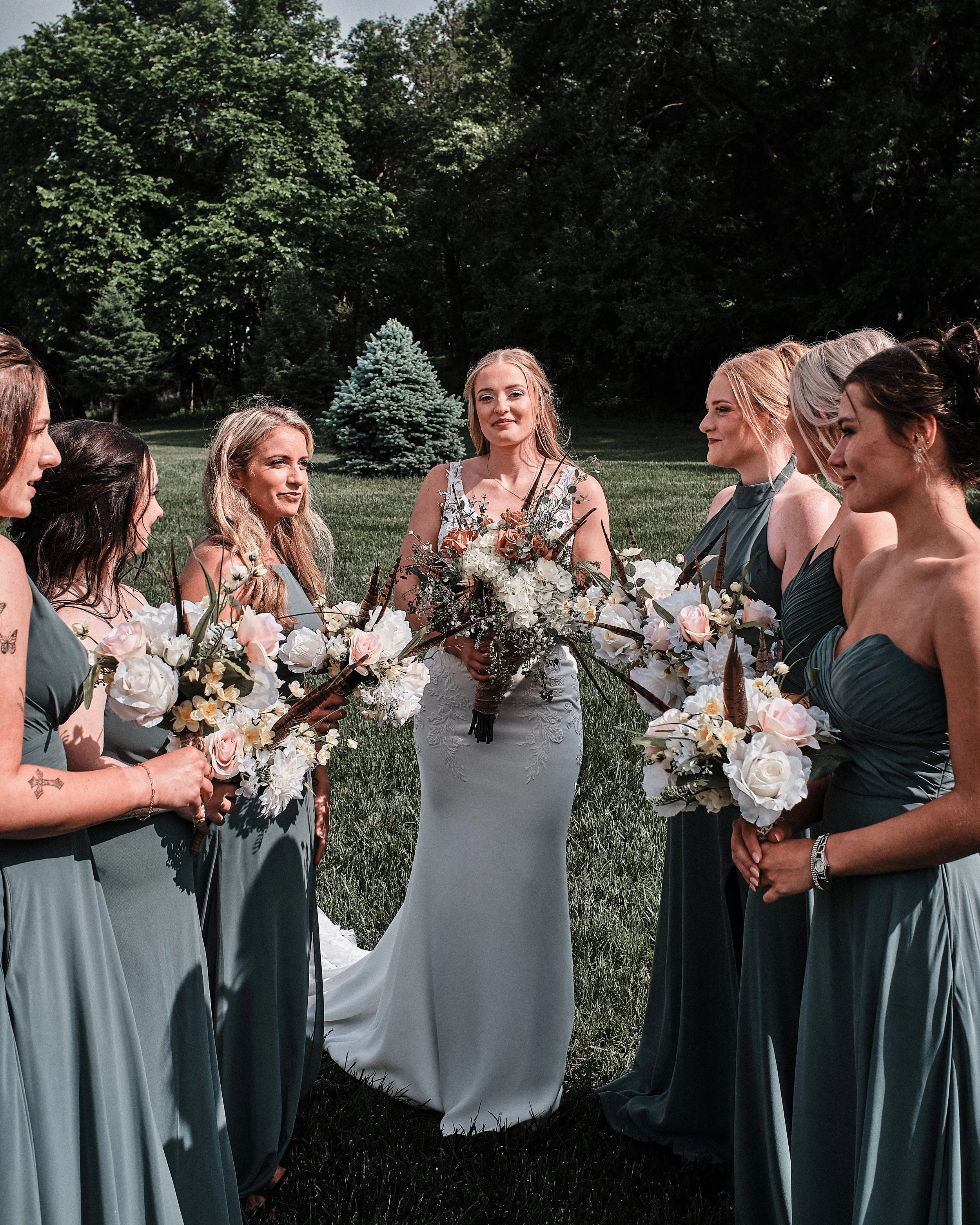 A bride and six bridesmaids stand outside on a grassy area, holding bouquets of white and blush flowers with greenery. The bride is wearing a white lace wedding gown and carrying a bouquet, with her hair styled in loose waves. The bridesmaids wear ma