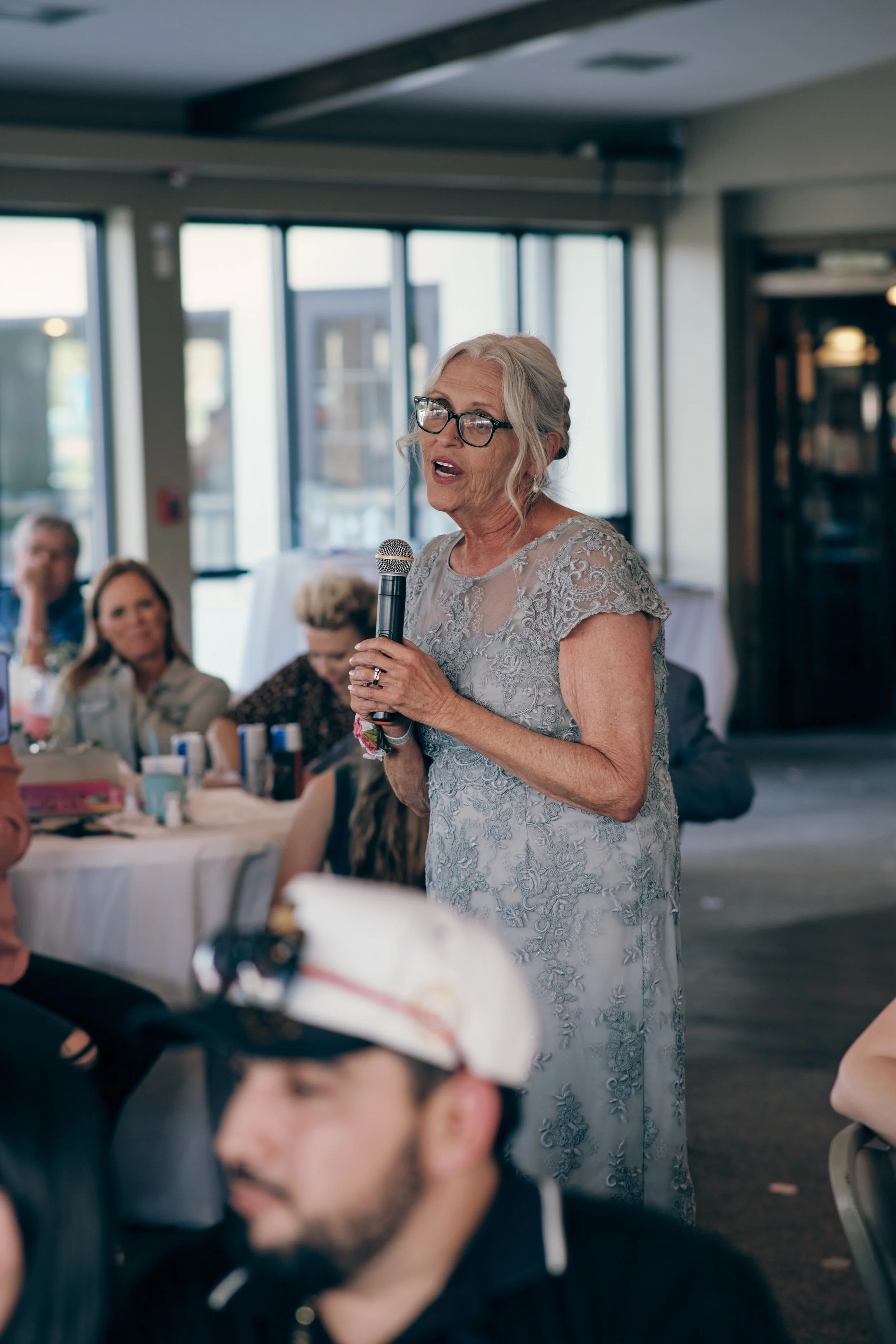 An elderly woman with gray hair and glasses speaking into a microphone at a formal gathering in a bright room with large windows. People are seated at round tables, listening. The woman is wearing a light-colored, embroidered dress.