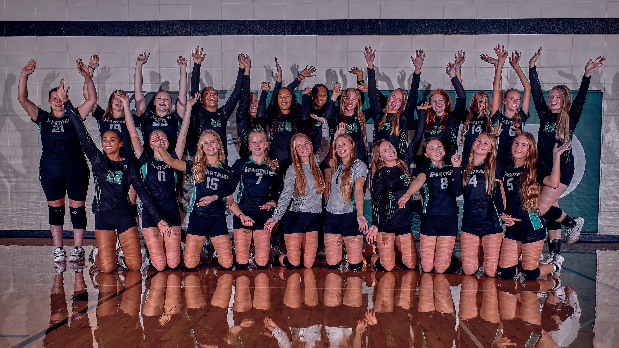 A group of female volleyball players wearing dark team uniforms with numbers and the word 'Spartans' on the front, posing in a gymnasium with a wooden floor and a tiled wall behind them. They are smiling, some raising their hands, with some kneeling in the front row.