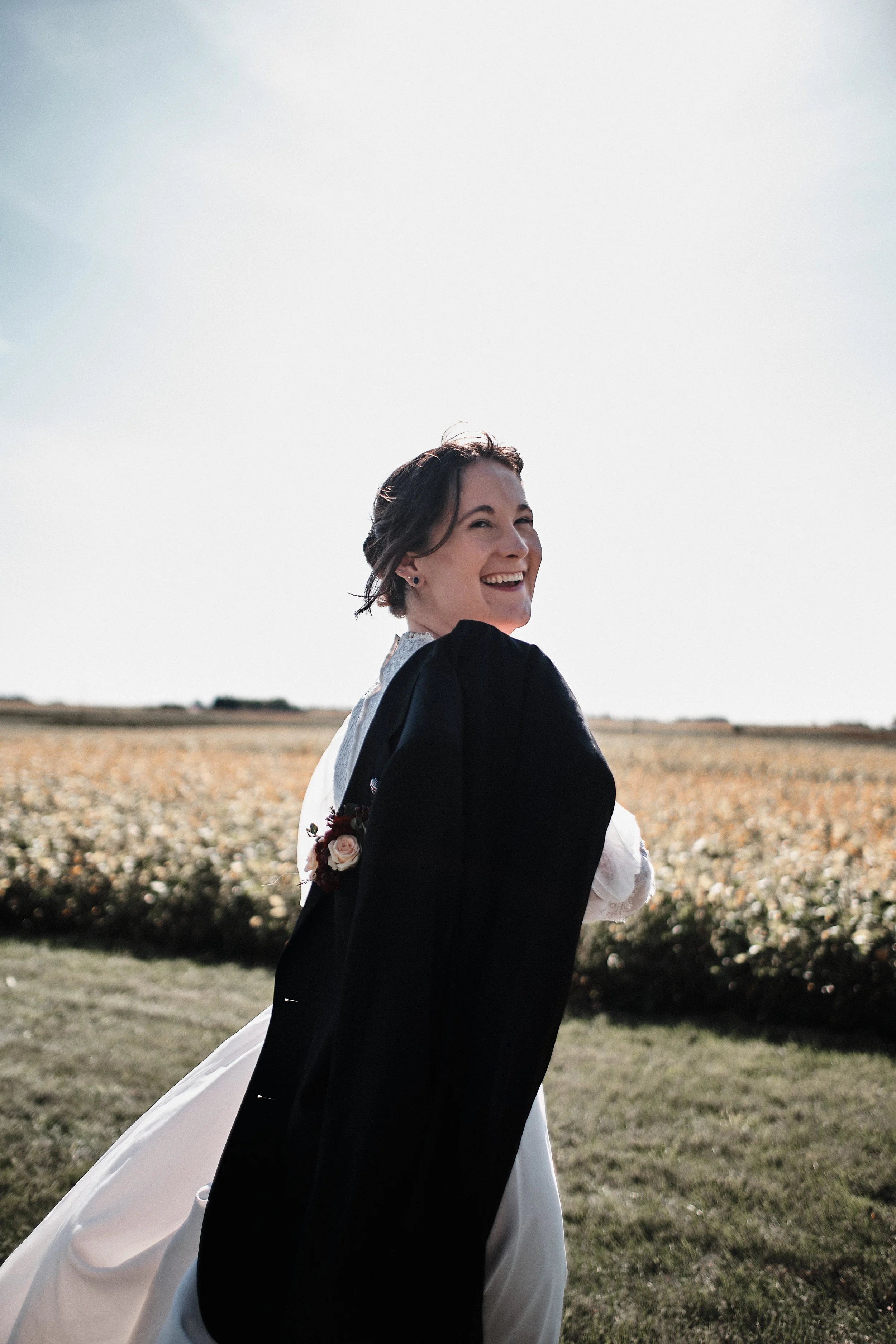A woman in a black and white wedding dress standing outdoors in a field, smiling, with a blue sky and distant trees in the background.