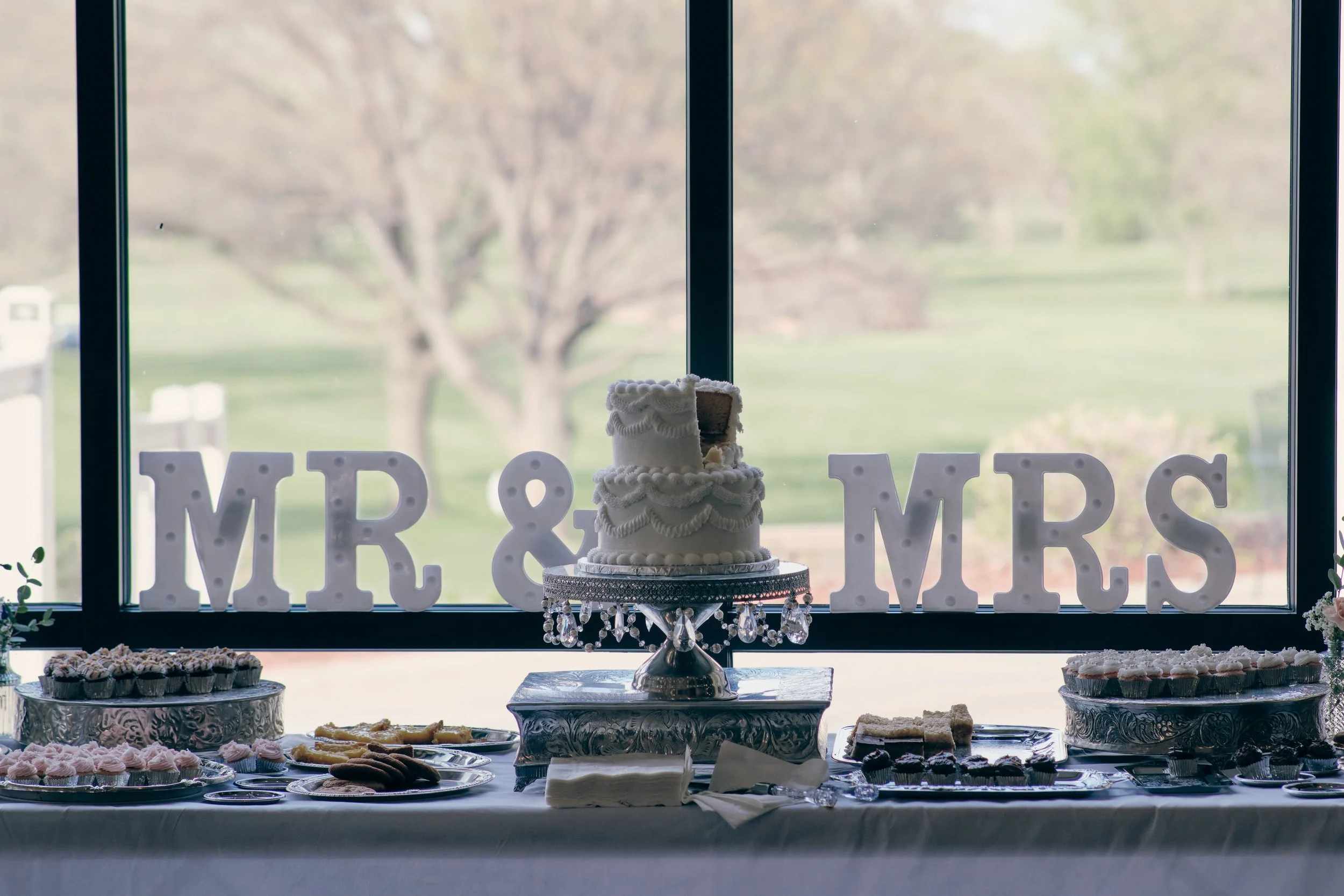 A dessert table with a decorated tiered cake on a silver stand in the center, surrounded by various cupcakes and cookies, with white illuminated letters spelling 'MR & MRS' in the background and a window showing an outdoor view.