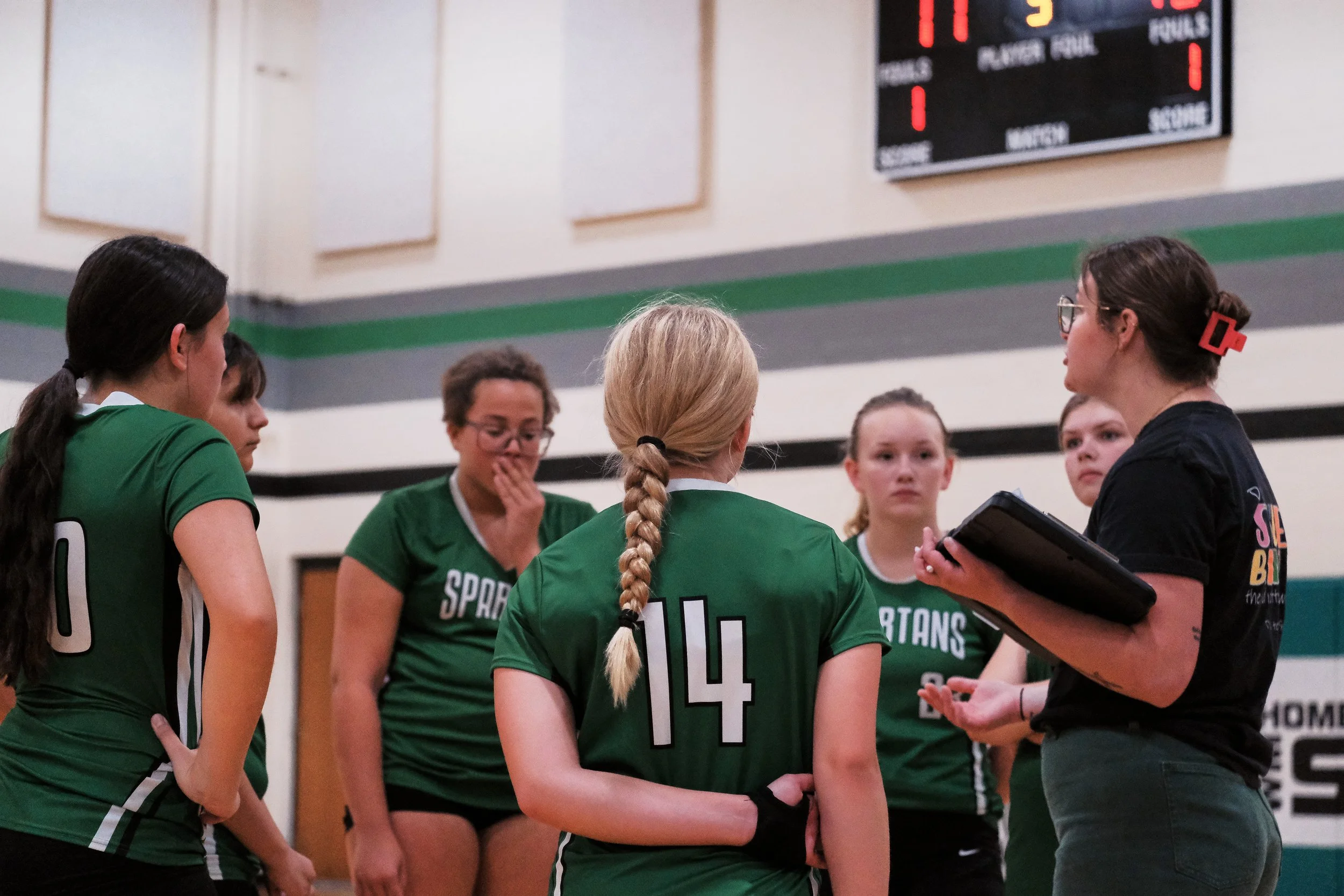 A group of young female volleyball players in green jerseys with white and black accents, gathered around their coach in a gymnasium. The players are listening intently as the coach speaks, holding a clipboard, with a scoreboard visible in the background.