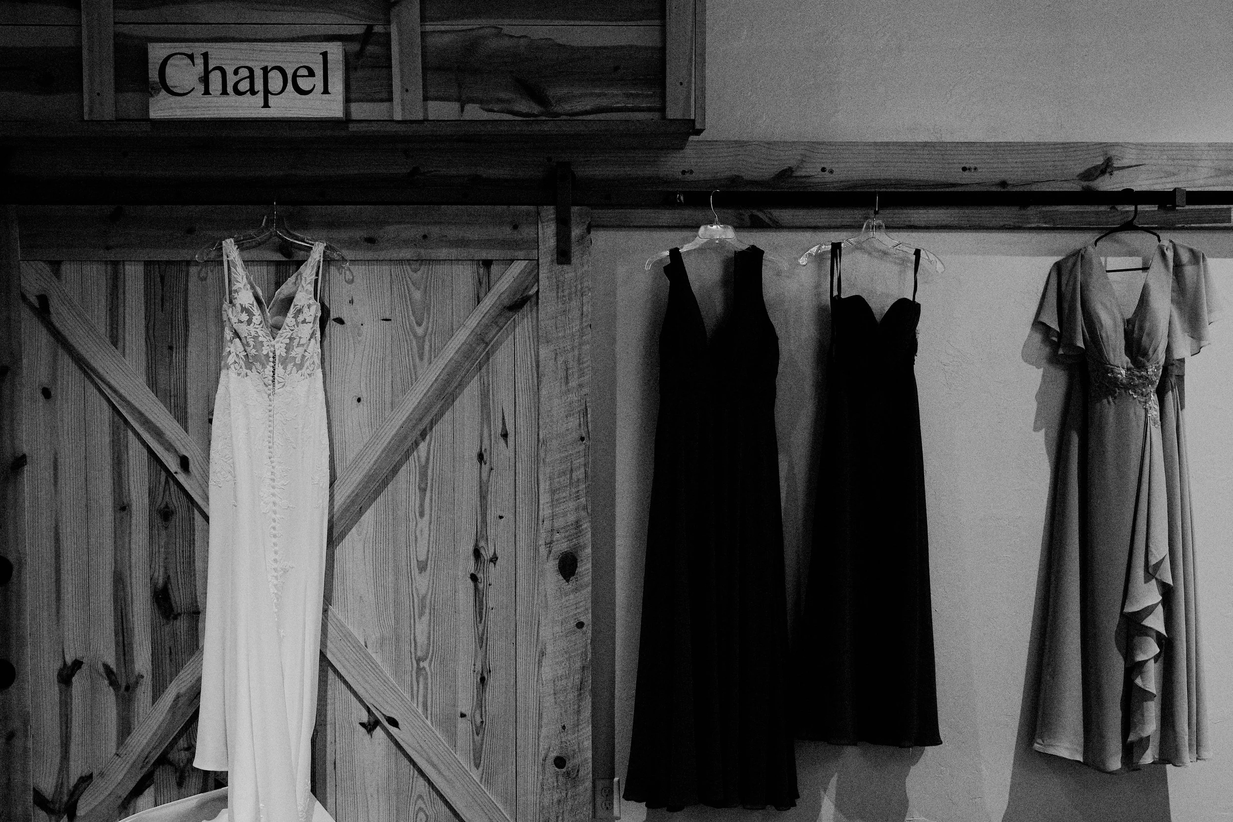 Black and white photo of dresses hanging on a horse rail in a wooden room with a sign that reads "Chapel".