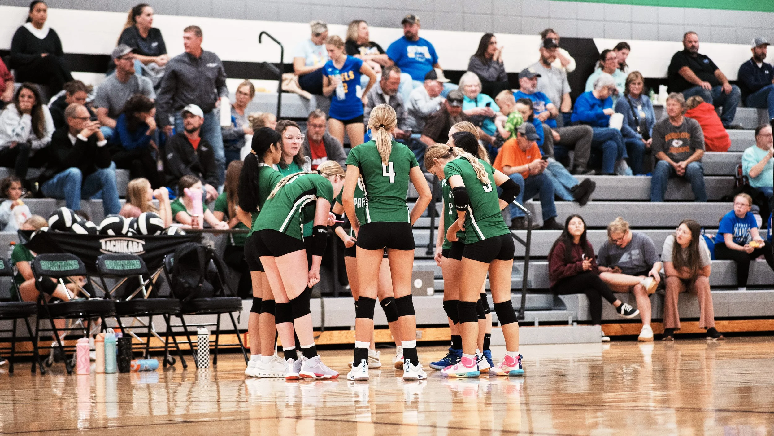 A volleyball team in green uniforms gathered in a huddle during a game in an indoor gymnasium, with spectators seated in the bleachers watching.