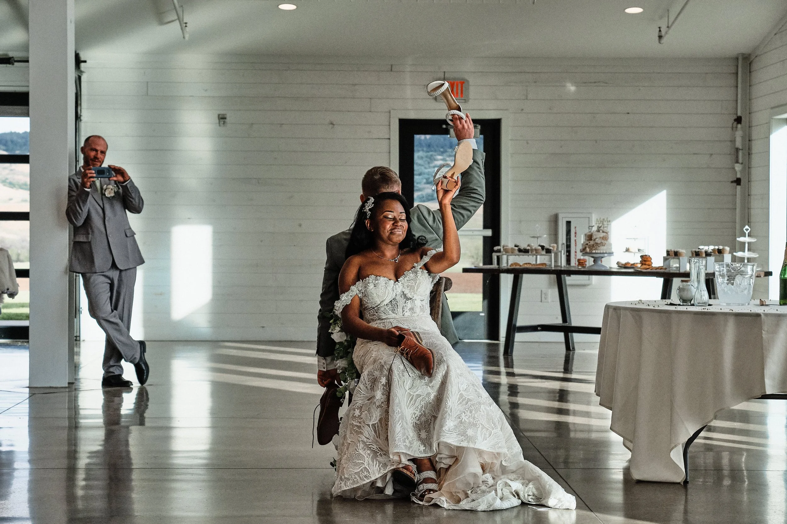A bride sitting on a chair with a big smile, holding her dress as a dancer with a sandal on her hand performs a dance behind her at a wedding reception. In the background, a man in a suit is taking a photo.