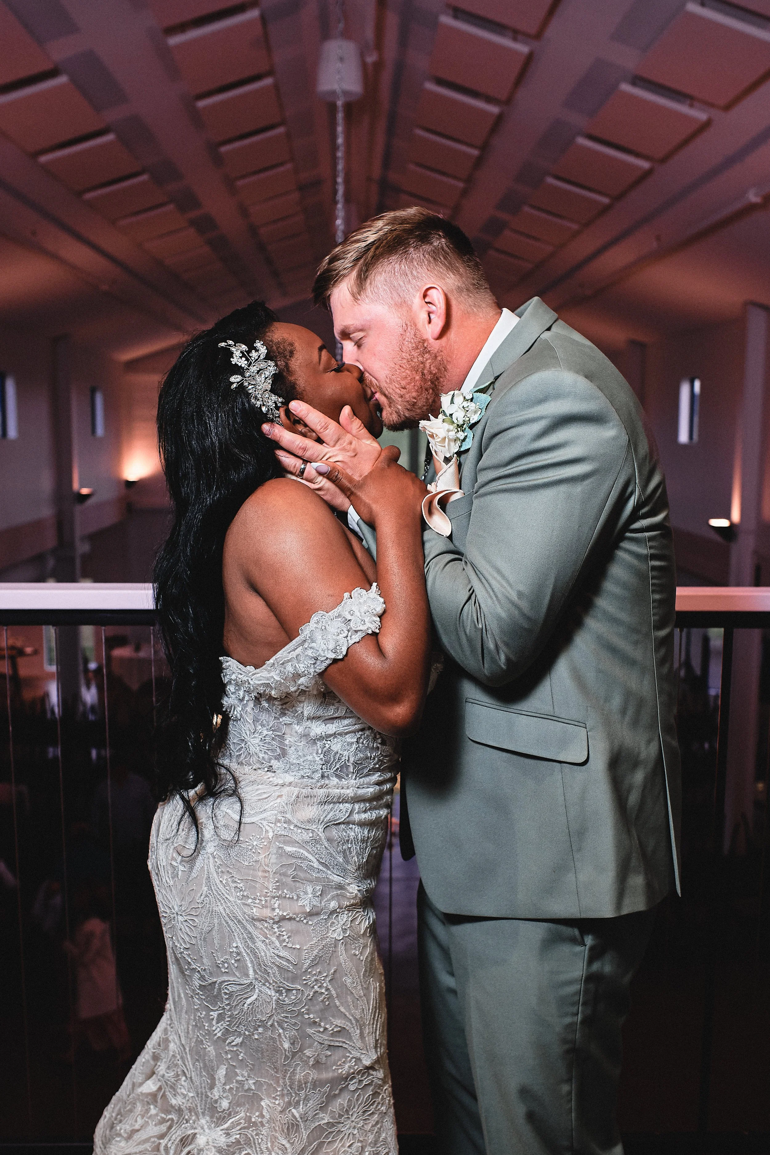 A couple, a woman in a lace wedding gown and a man in a gray suit, are sharing a kiss at a wedding reception. The woman has long dark hair decorated with a silver hairpiece, and the man has short hair and a beard, wearing a boutonniere with white flo
