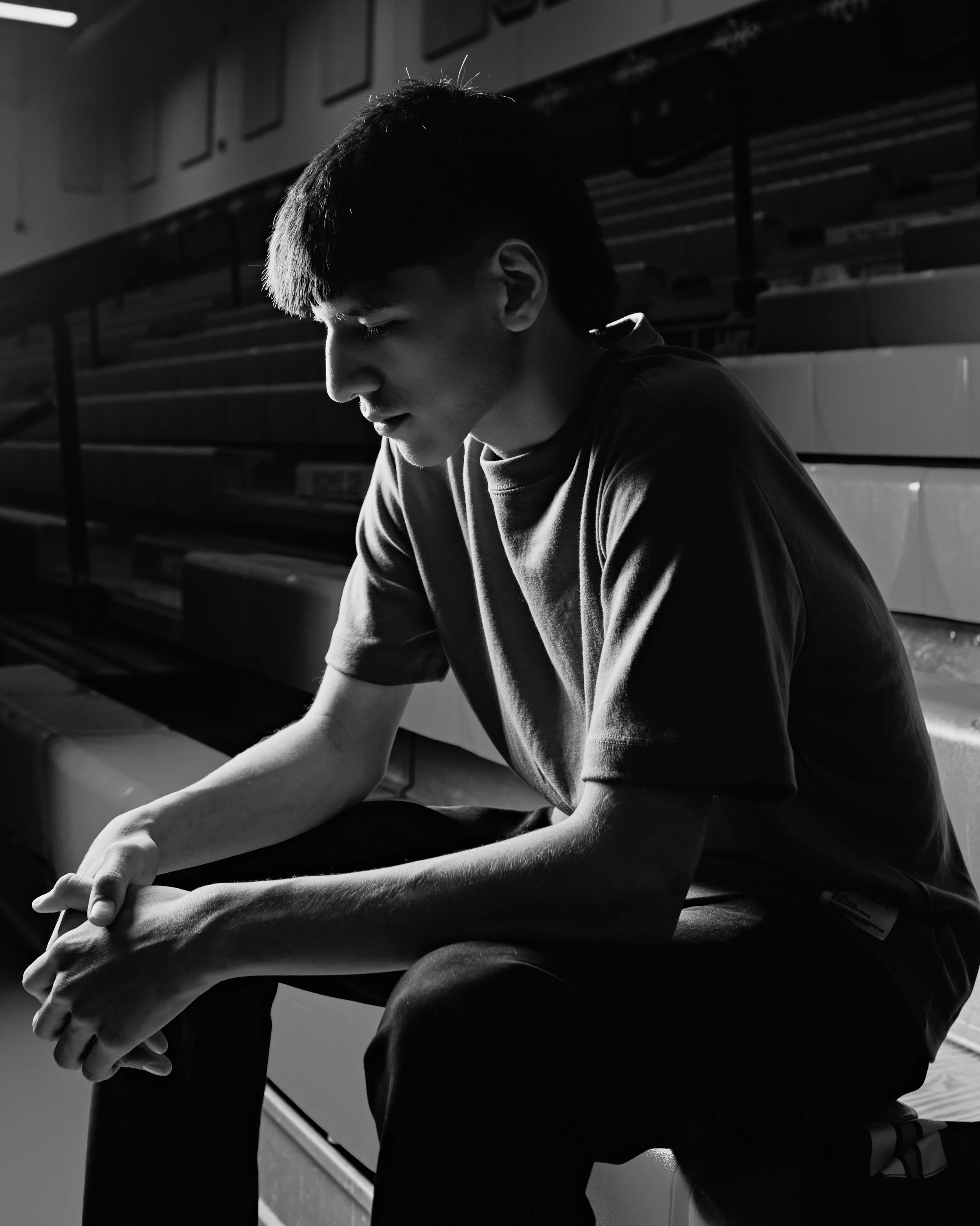 A young man with short hair sitting on a basketball court, leaning forward, with a serious expression. He is wearing a T-shirt and shorts. The background has empty bleacher seats.