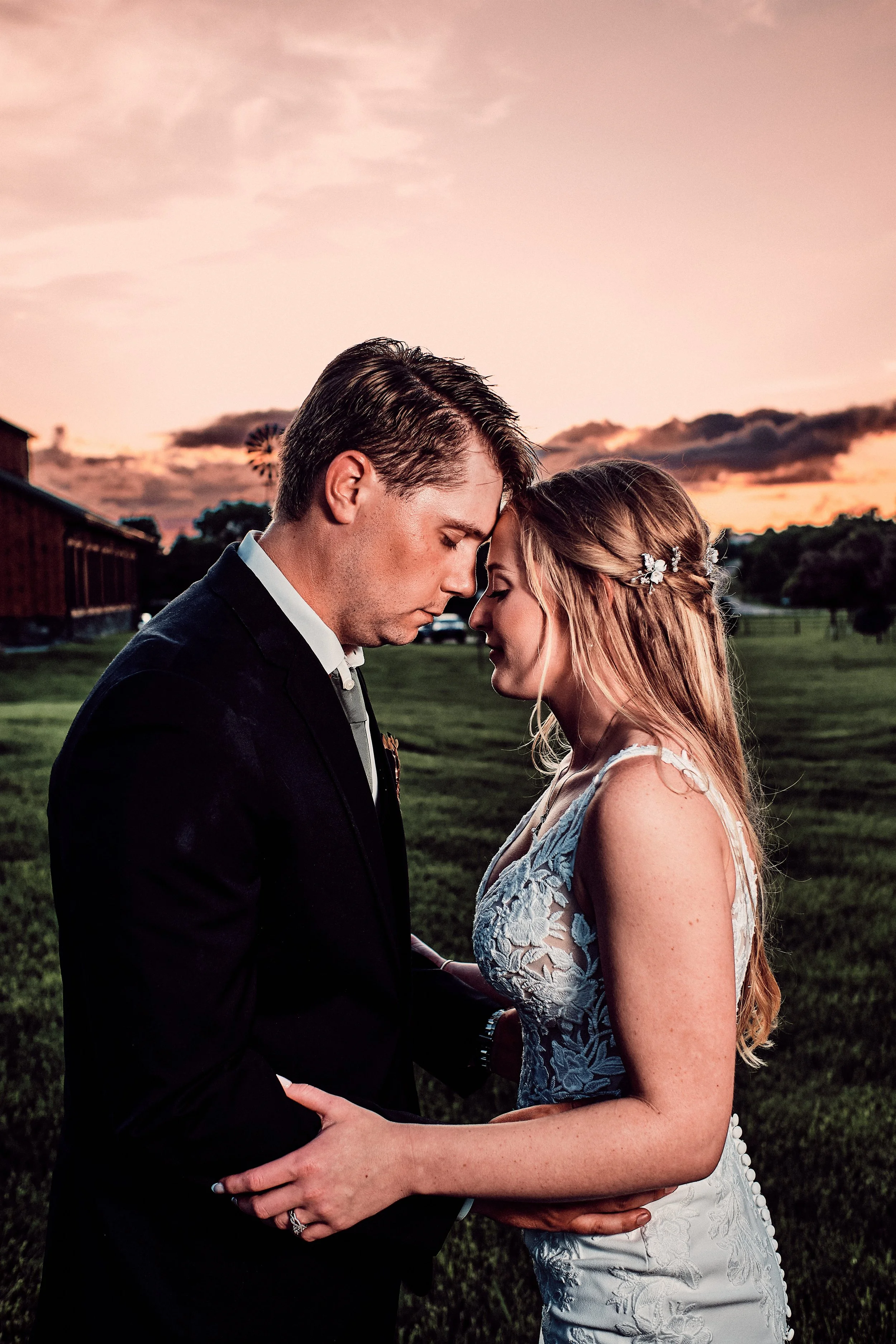 A bride and groom touching foreheads and holding hands outdoors at sunset, with a barn and a windmill in the background, during their wedding.