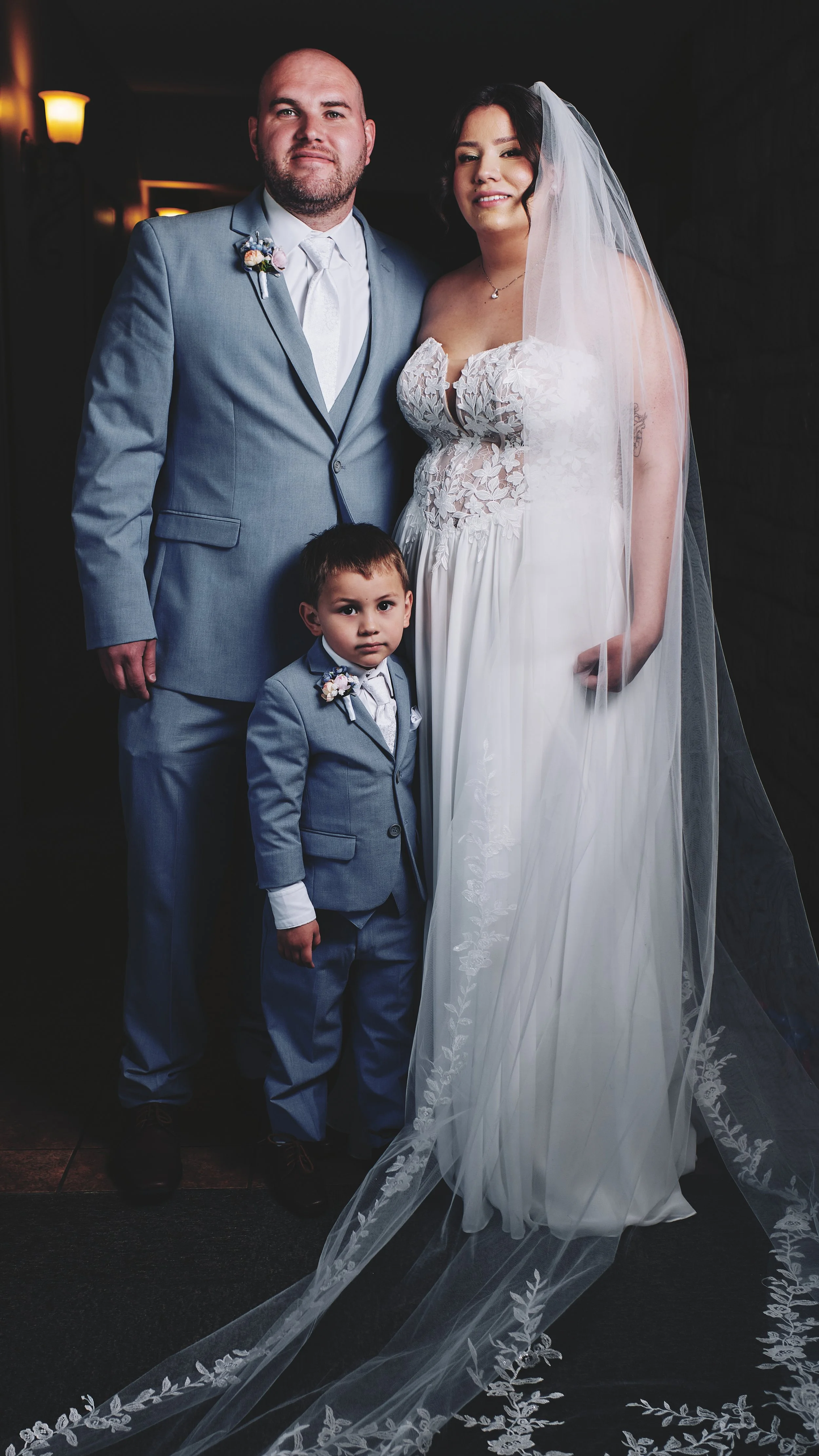 A wedding photo of a man, woman, and young boy, all dressed in formal attire, standing indoors with dim lighting.