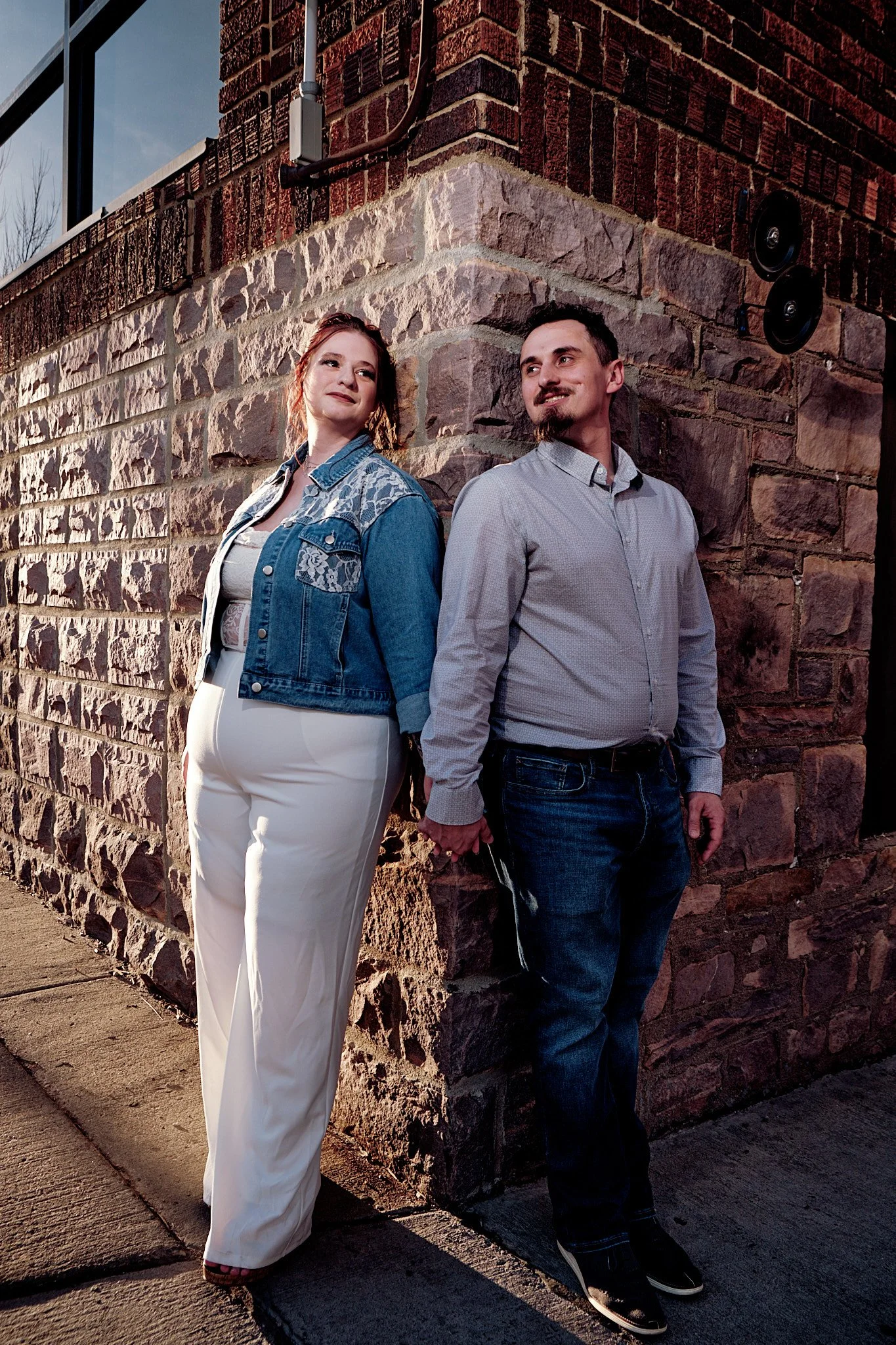A man and woman stand back-to-back against a brick wall during sunset. The woman is wearing a denim jacket with lace details and white pants, while the man is dressed in a light gray shirt and dark jeans. They are holding hands behind their backs and