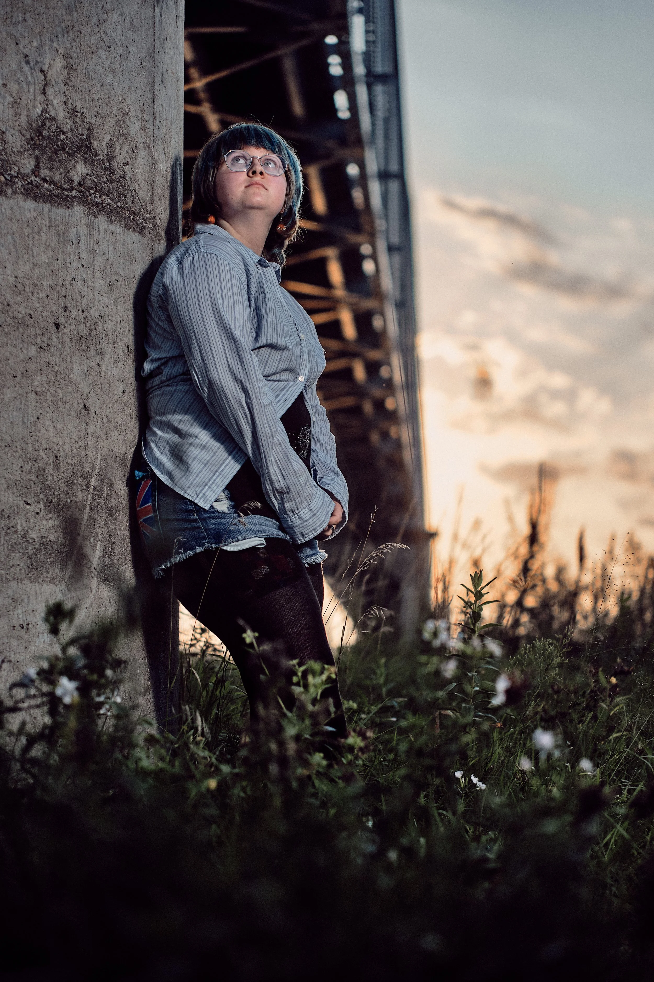 A young person with glasses and blue hair, wearing a striped shirt and ripped jeans, leaning against a concrete wall outdoors at sunset, looking upward.