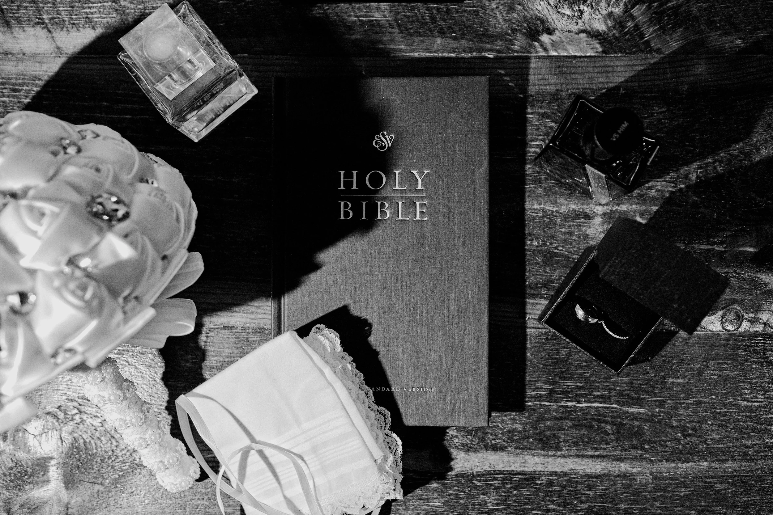 A flat lay of a holy bible, perfume bottles, a gift bag, and jewelry on a wooden surface in black and white.