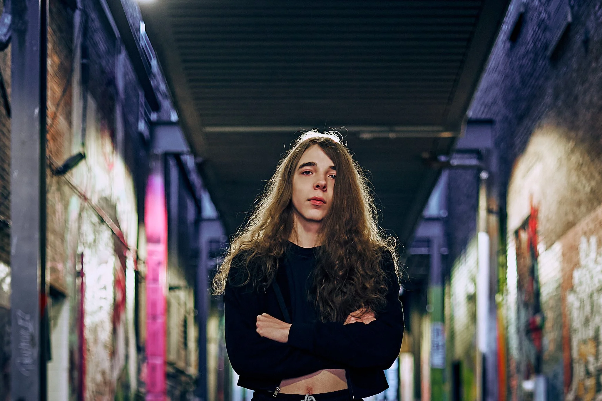 A young woman with long curly hair, arms crossed, standing in an alleyway between colorful graffiti walls.