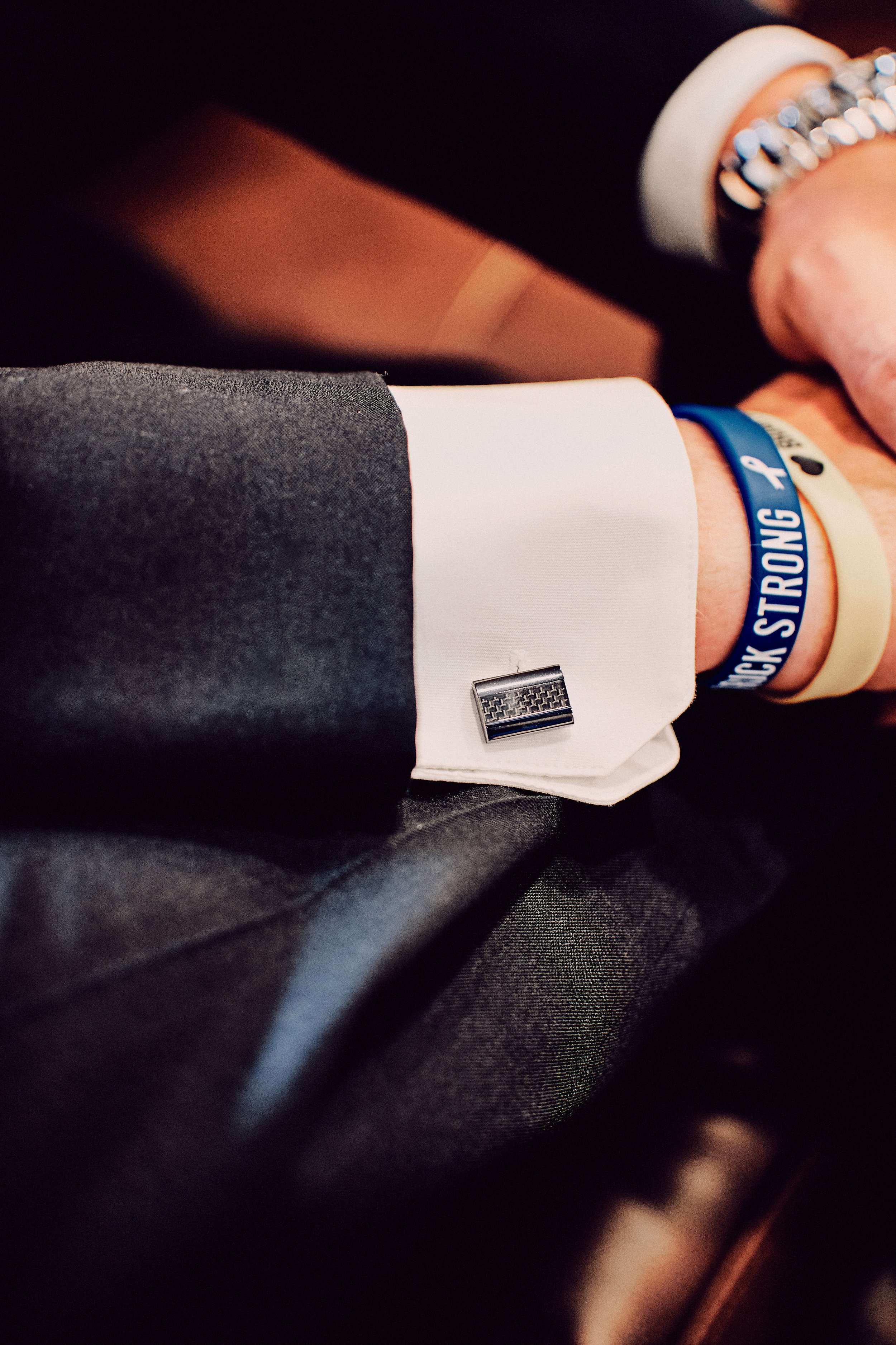 Close-up of a person's hand wearing a wristband that reads "NECK STRONG" on a black suit, white shirt, and cufflink, with a blurred background.