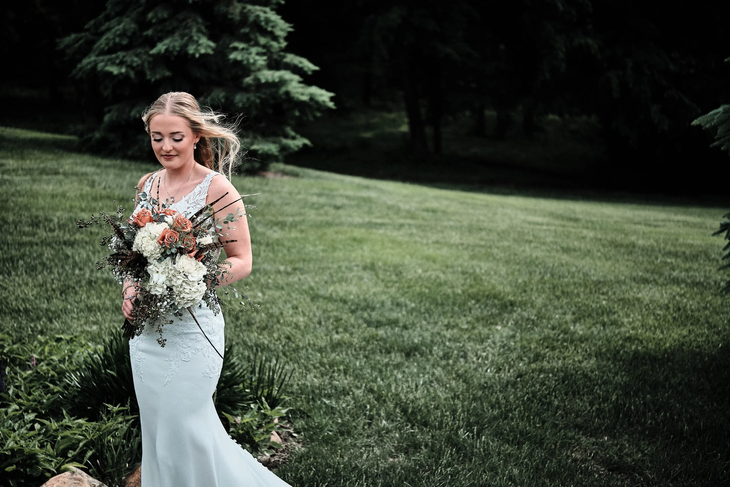 A bride standing outdoors on a lush green lawn, holding a bouquet of flowers, wearing a white lace wedding dress with floral embroidery and a sleeveless design, with her hair styled in loose waves.