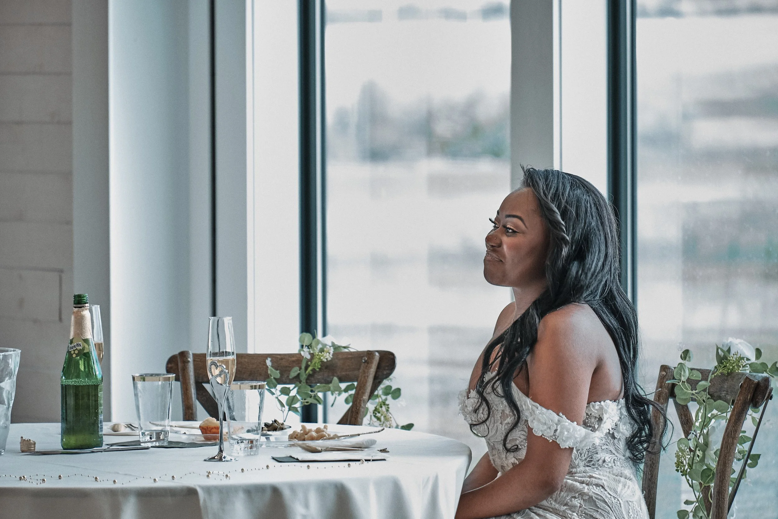 A woman with long dark hair wearing a white lace dress sitting at a dining table with wedding decor.