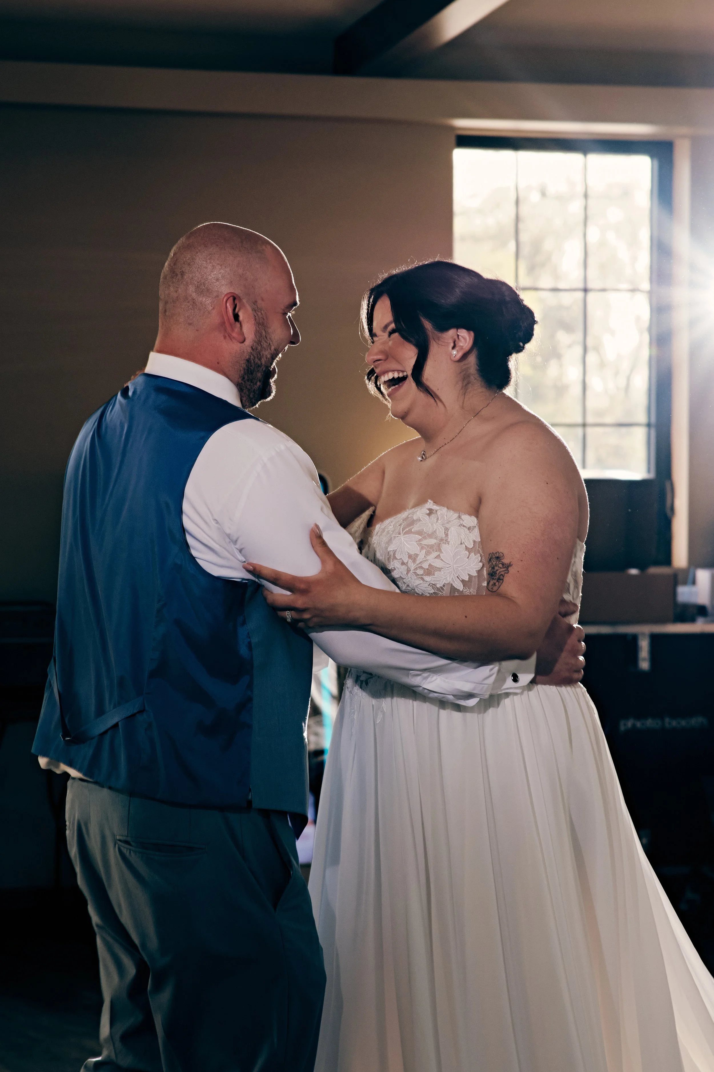 A couple dressed in wedding attire, smiling joyfully and sharing a dance indoors near a window with sunlight streaming in.