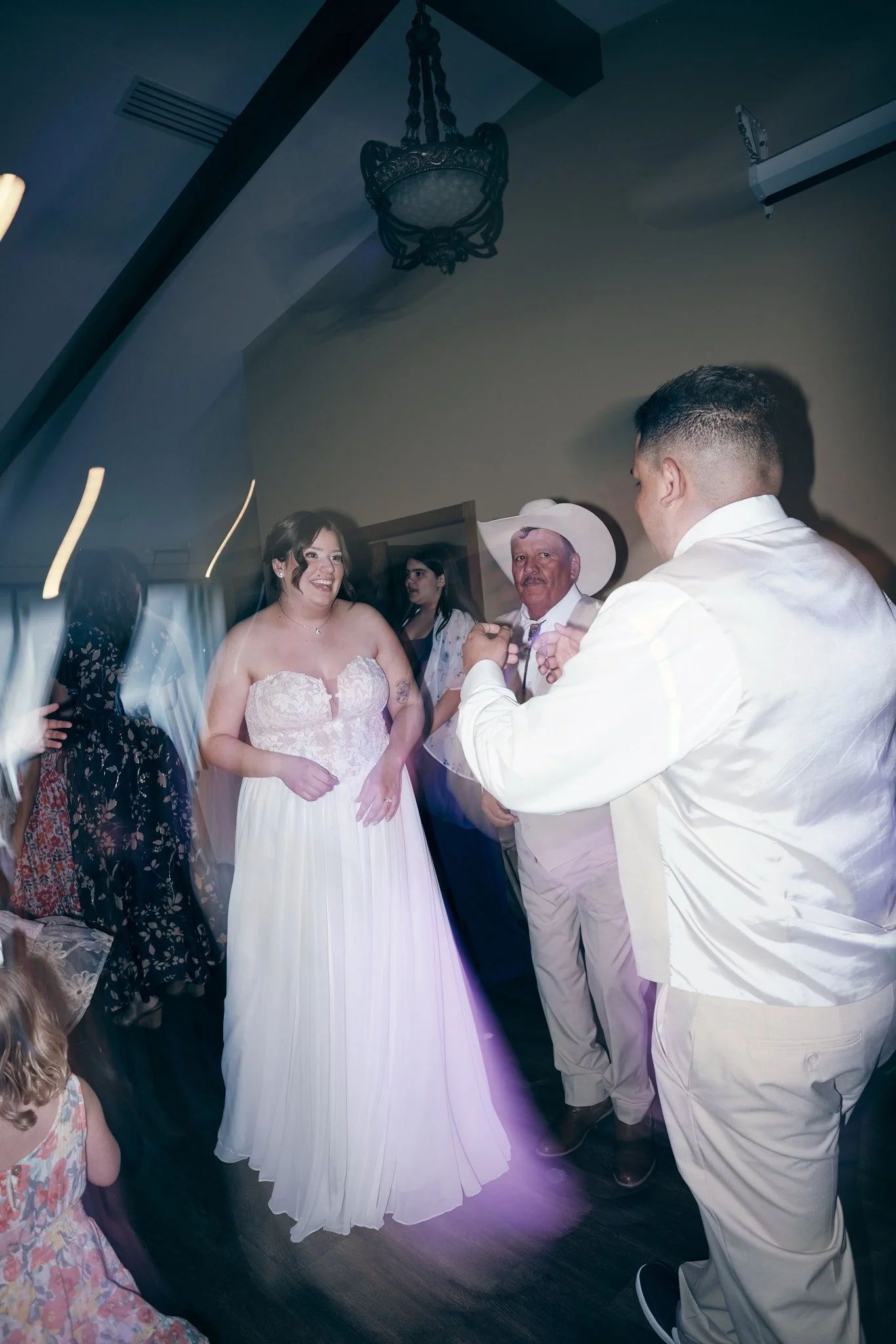 A bride in a white wedding dress smiles while dancing with her father at a wedding reception. Other guests are visible in the background.