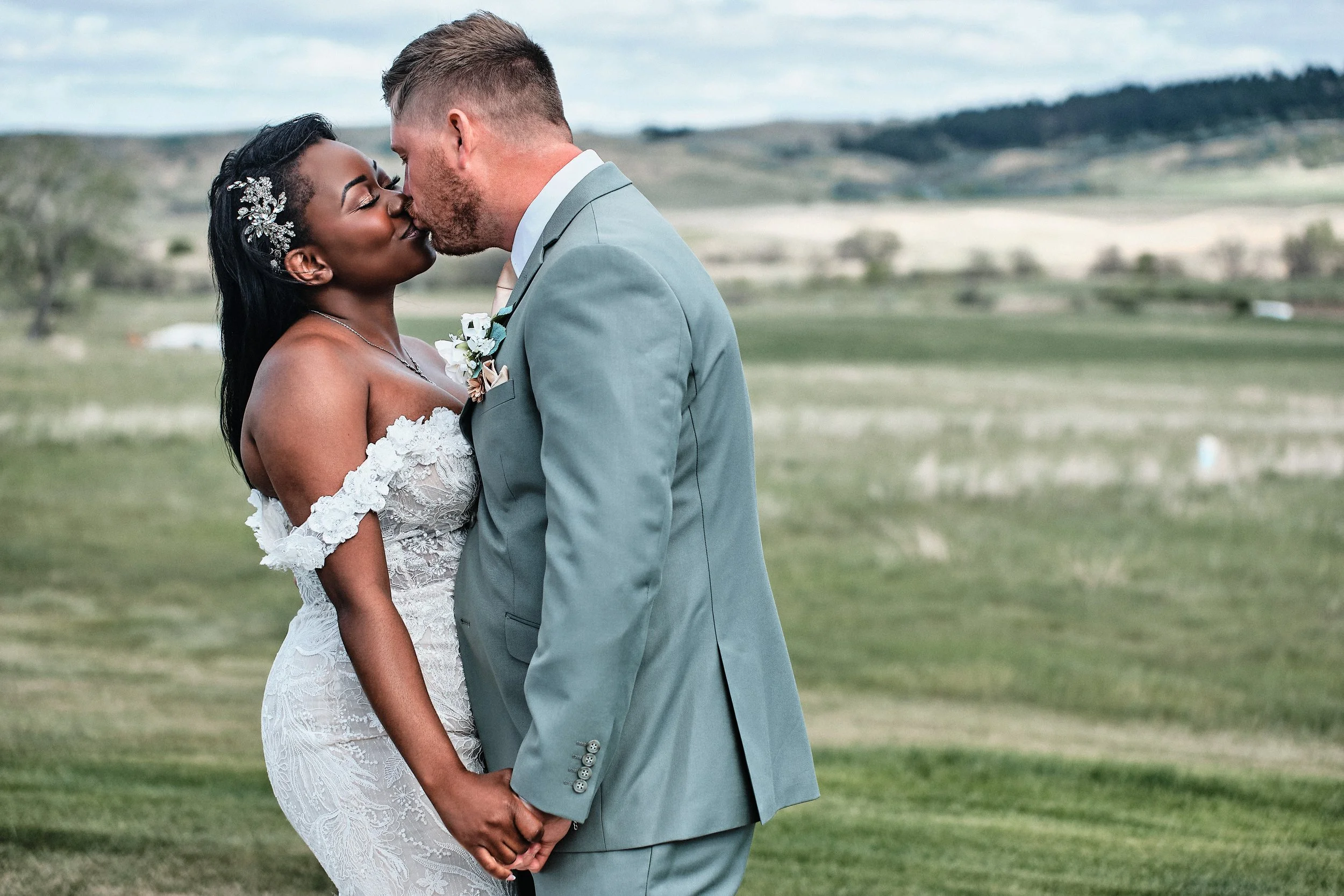 A newlywed couple standing outdoors, holding hands and kissing, with a landscape of open fields and rolling hills in the background.