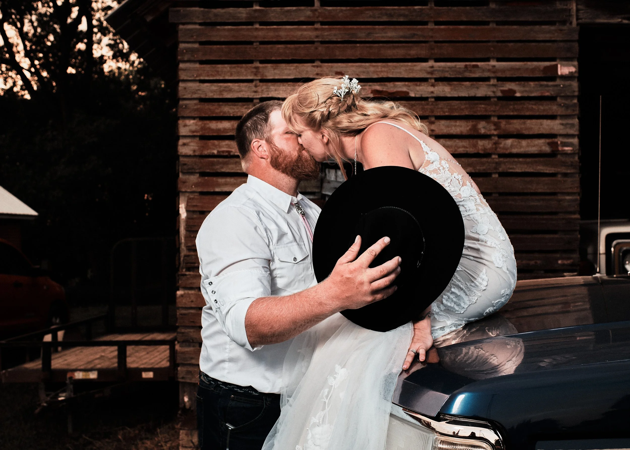 A couple shares a kiss at sunset. The woman is dressed in a white wedding gown with lace details, and the man is holding a black hat. They are standing in front of a rustic wooden wall, next to a silver car.