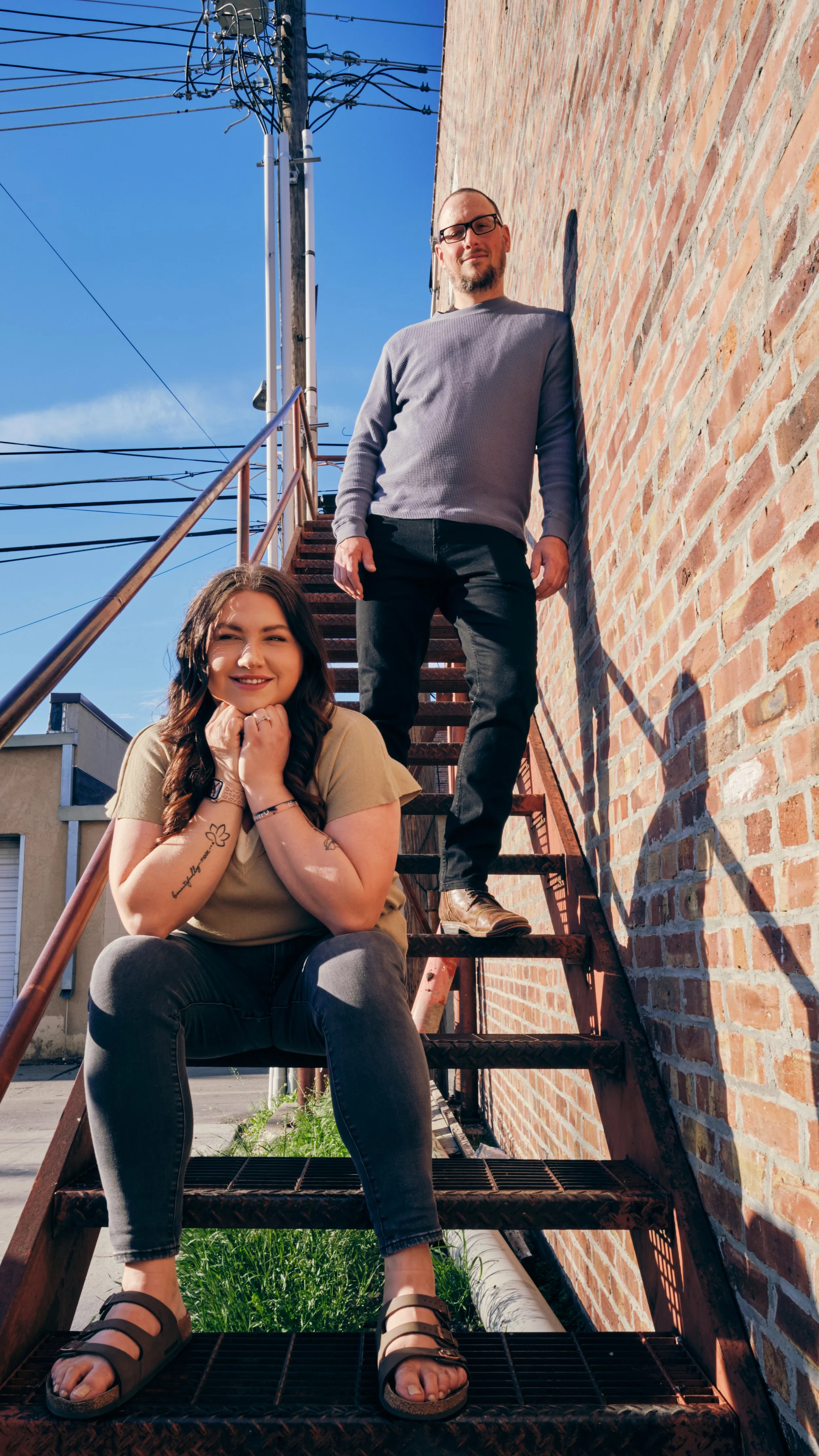 A woman sitting on an outdoor metal staircase, smiling, and a man standing behind her, leaning against a brick wall, both in casual clothing under a clear blue sky.
