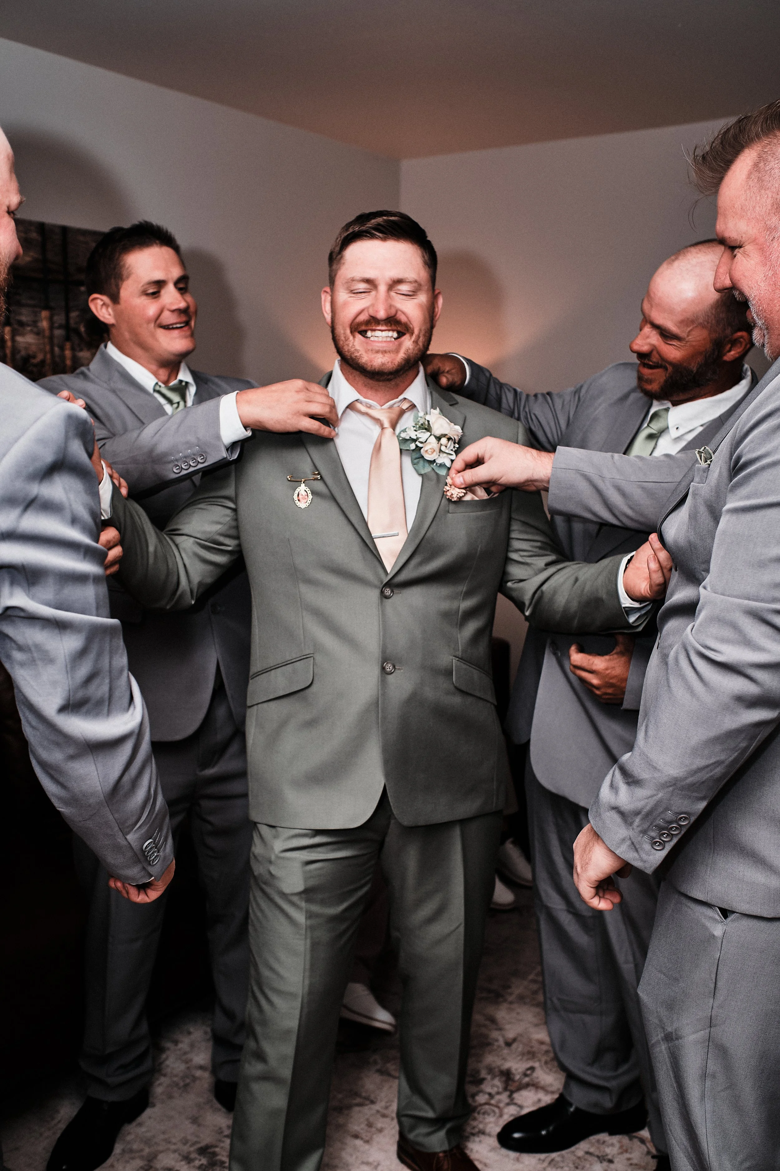 A groom in a gray suit smiles with eyes closed while surrounded by five men in suits, who are helping him adjust his boutonniere during a wedding preparation.
