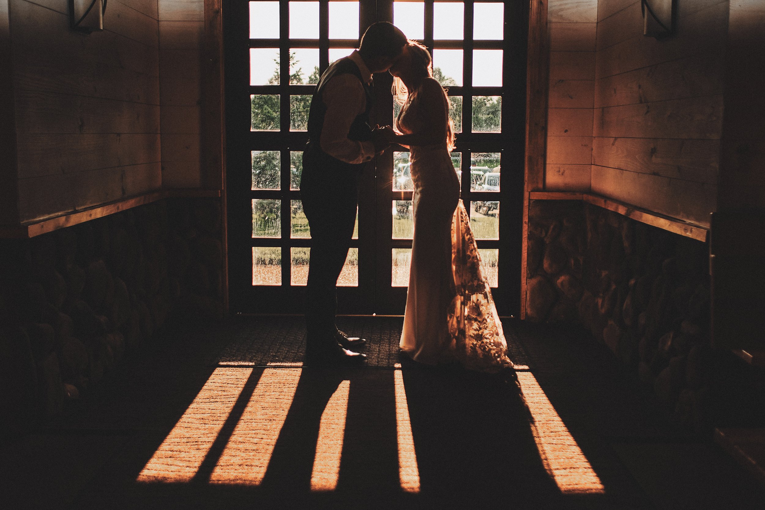 Silhouette of a couple standing close together inside a wooden building, with their foreheads touching, holding hands, backlit by sunlight coming through a large window with multiple panes, casting long shadows on the floor.