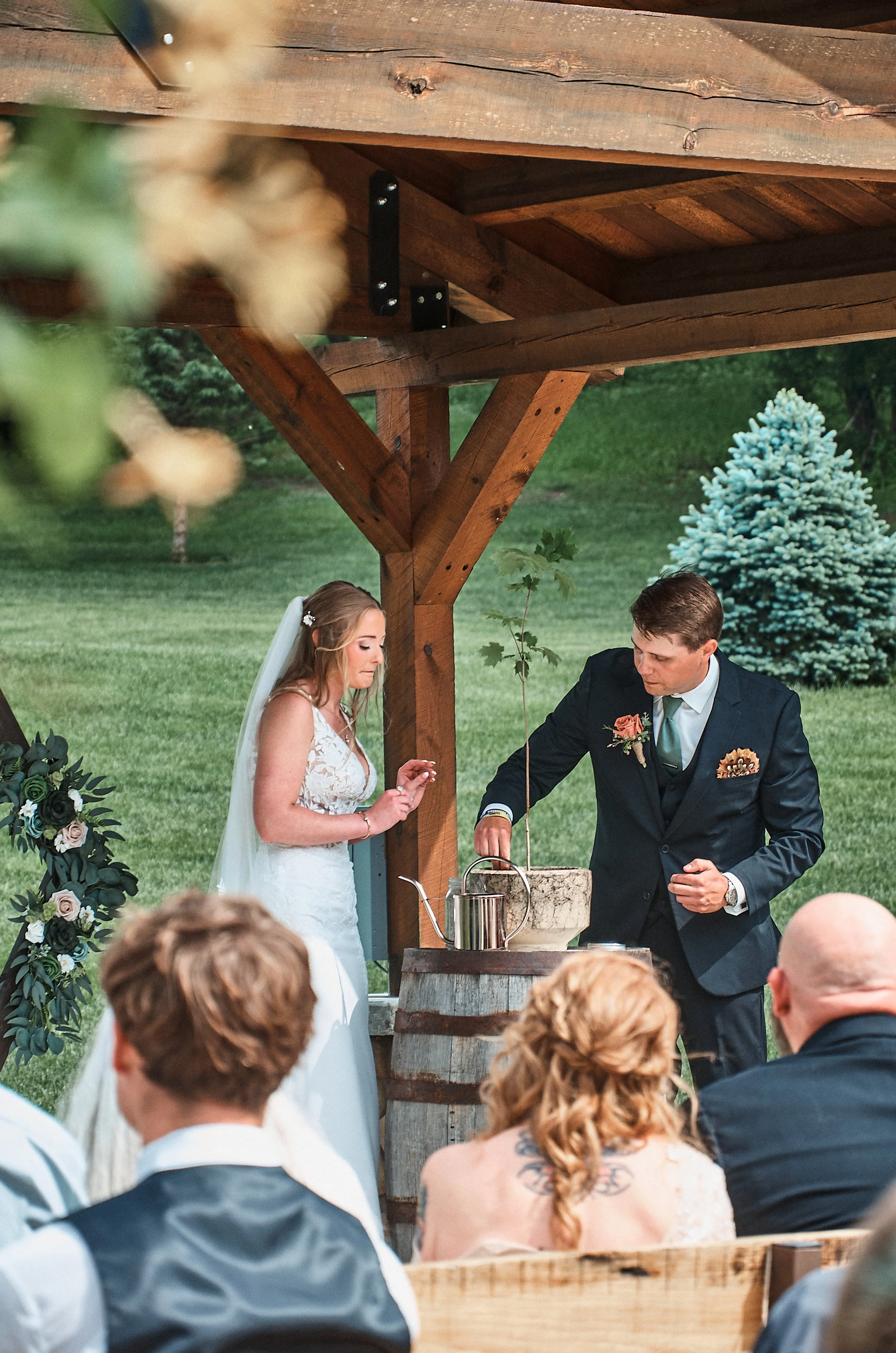 Bride and groom at an outdoor wedding ceremony standing under a wooden pavilion with guests seated in front of them.
