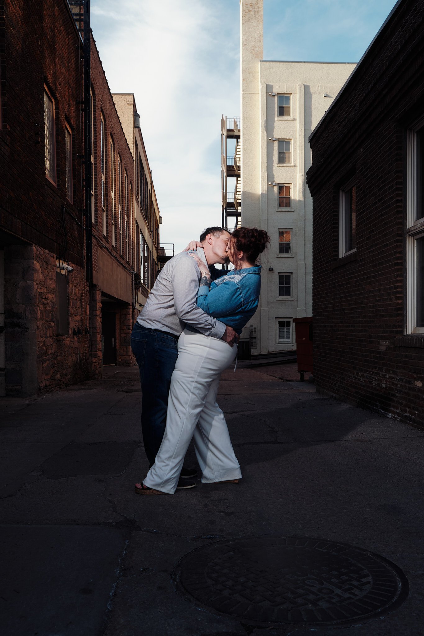 A couple sharing a kiss in an urban alleyway during daylight, surrounded by brick and white buildings.