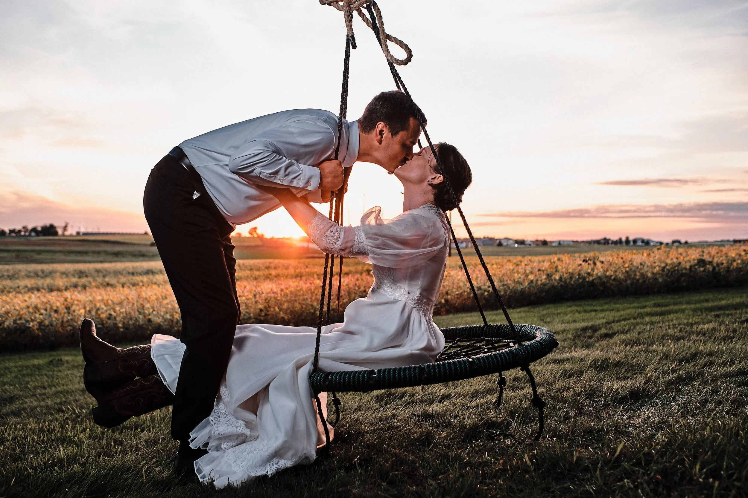 A man kneeling on a swing kissing a woman who is sitting on the swing with her eyes closed, during sunset in a field.