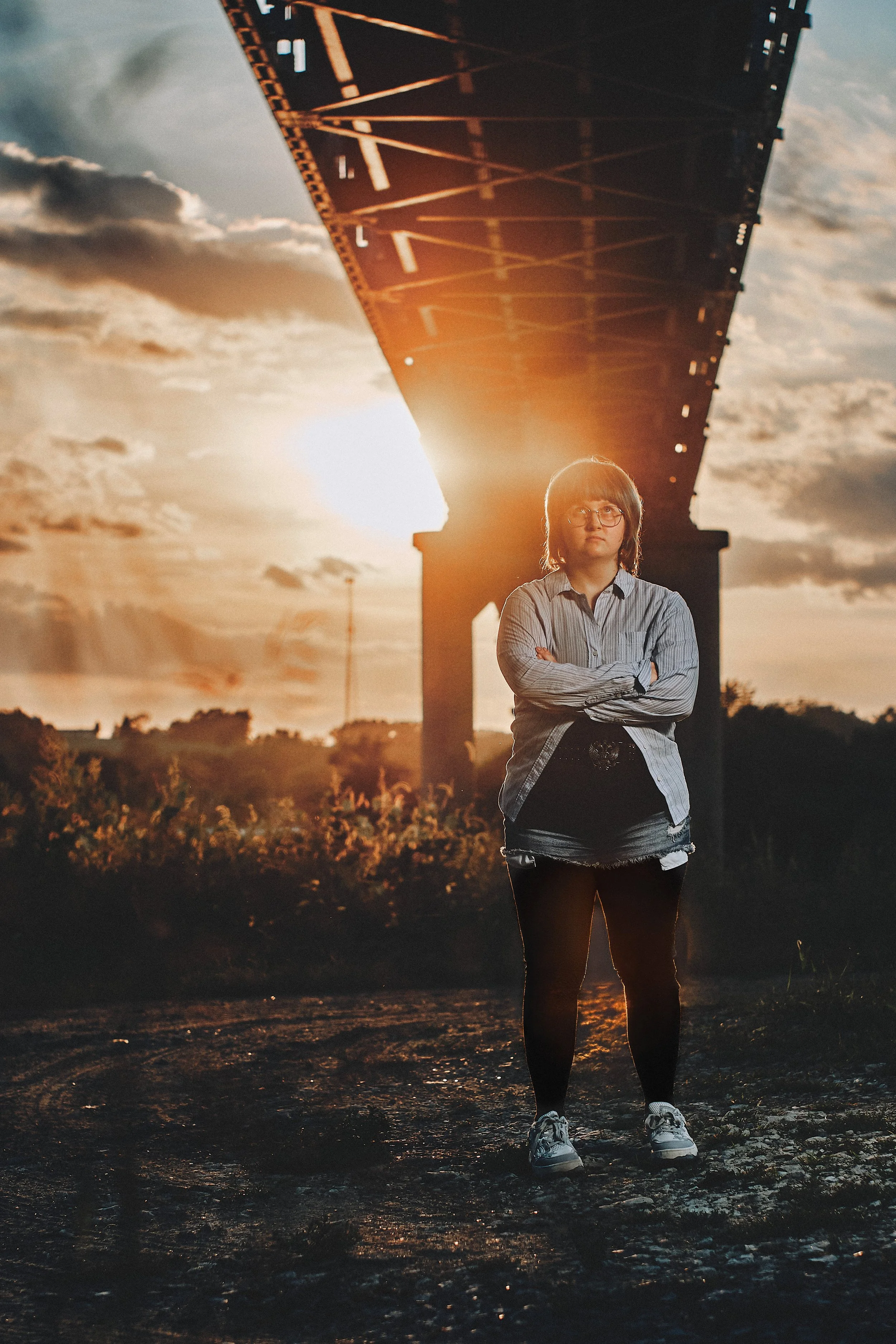 A person with glasses and a striped shirt stands with arms crossed under a bridge during sunset, with clouds and the sun in the background.