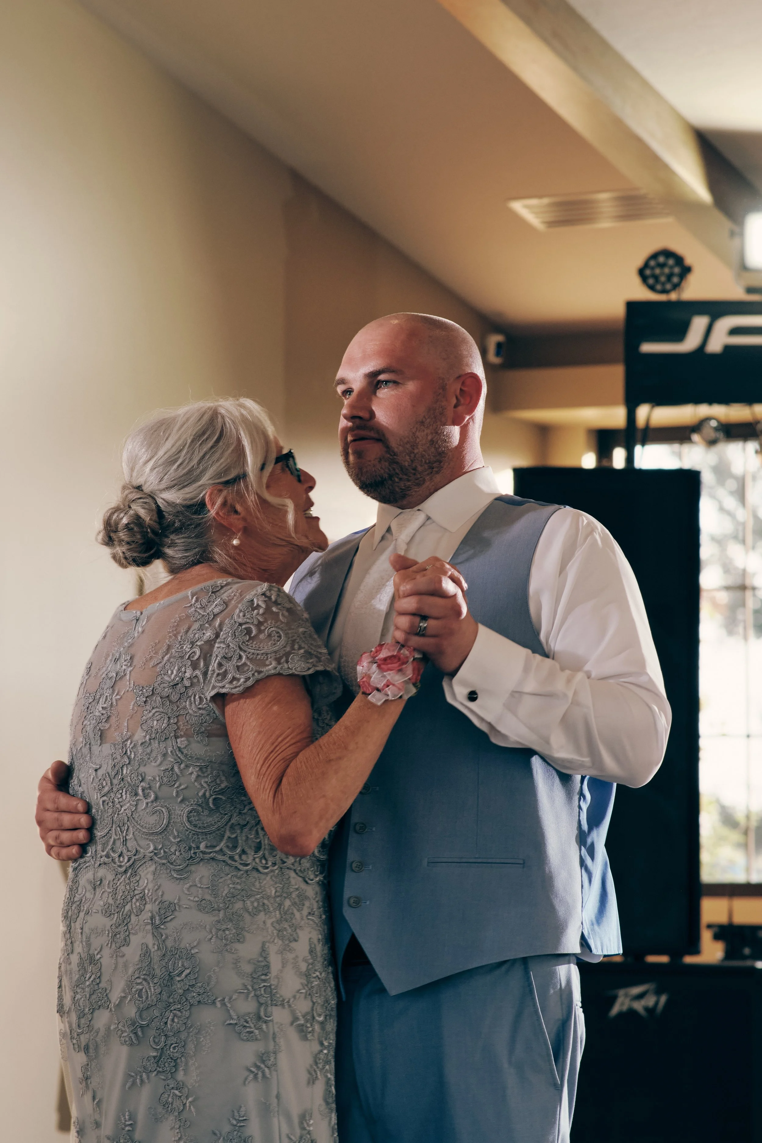 An elderly woman and a middle-aged man dance closely together at a wedding reception. The woman wears a lace dress and glasses, while the man wears a light blue vest and white shirt. They are dancing against a beige wall with windows and curtains in 
