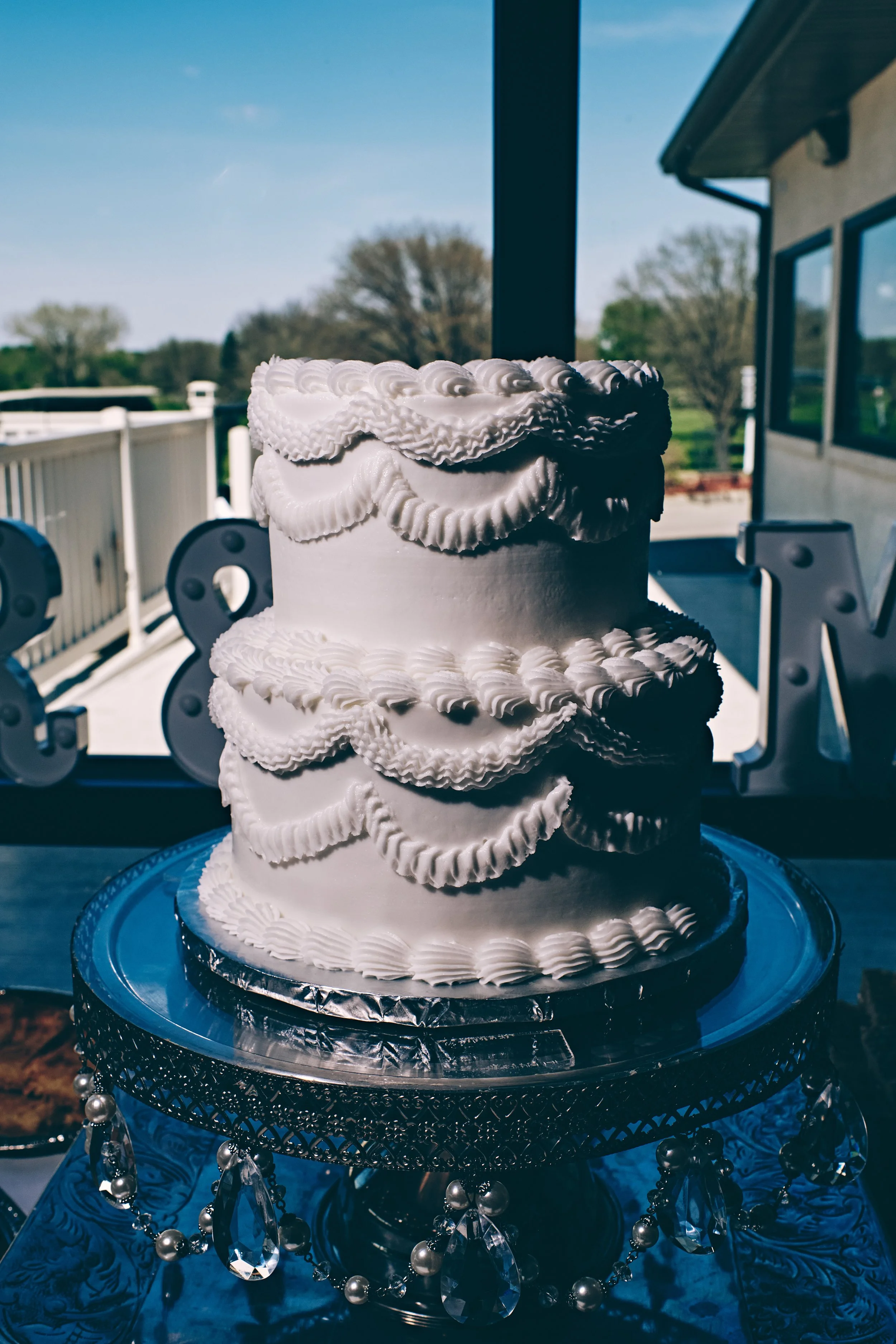 A white tiered cake with rosette and shell piping decoration on a silver cake stand, outdoors on a table with a blue patterned tablecloth, with a background of trees and a house.