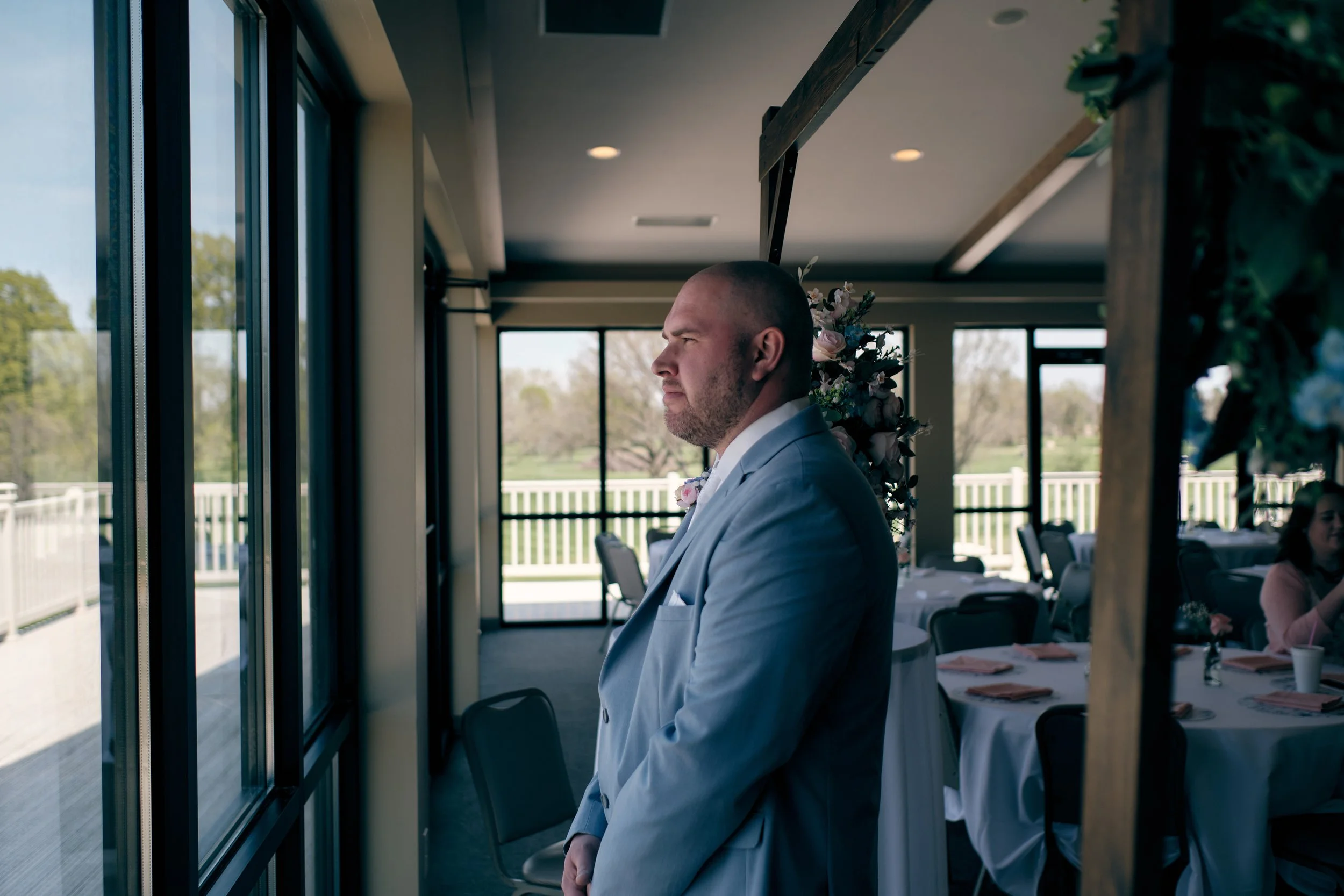 A man in a light blue suit looking out a window in a decorated room, with a floral arrangement behind him and other people sitting at tables in the background.