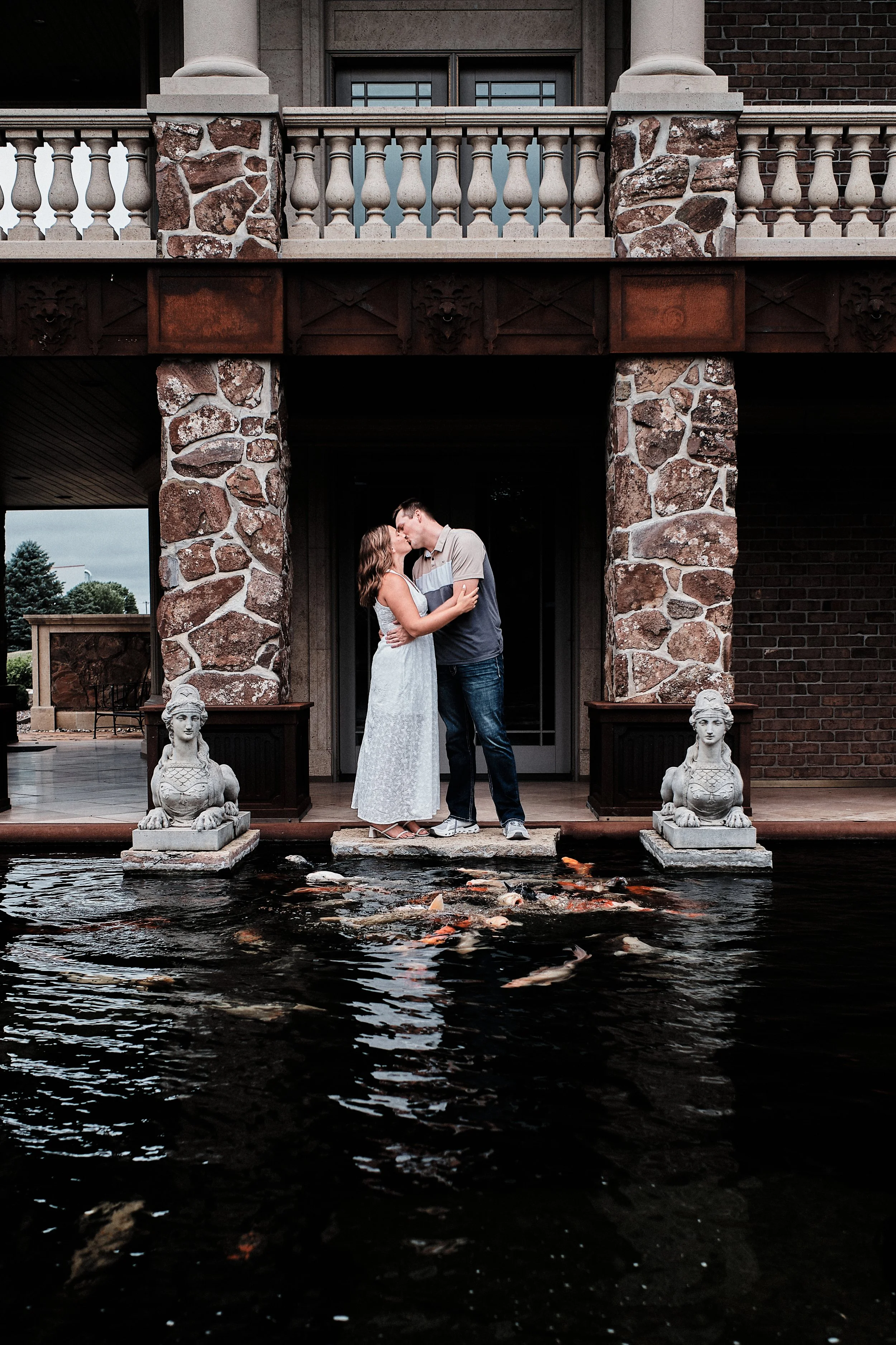 A couple is standing on a small stone platform in front of a pond, sharing a kiss. The couple is positioned under a stone archway with decorative pillars, and two stone statues of mythical creatures are on either side of the pond.