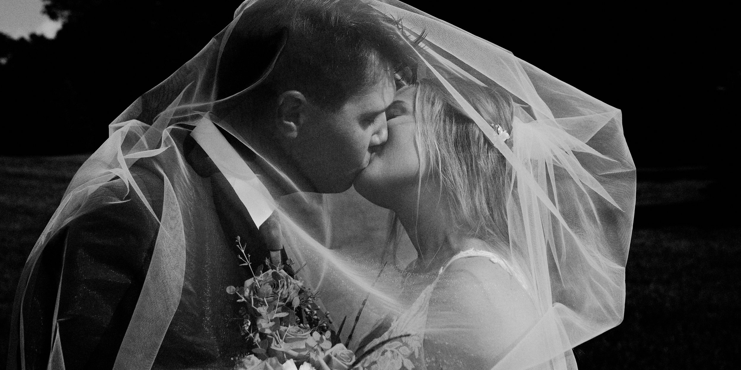 Black and white photo of a bride and groom kissing under a veil, with the groom holding a bouquet of flowers.
