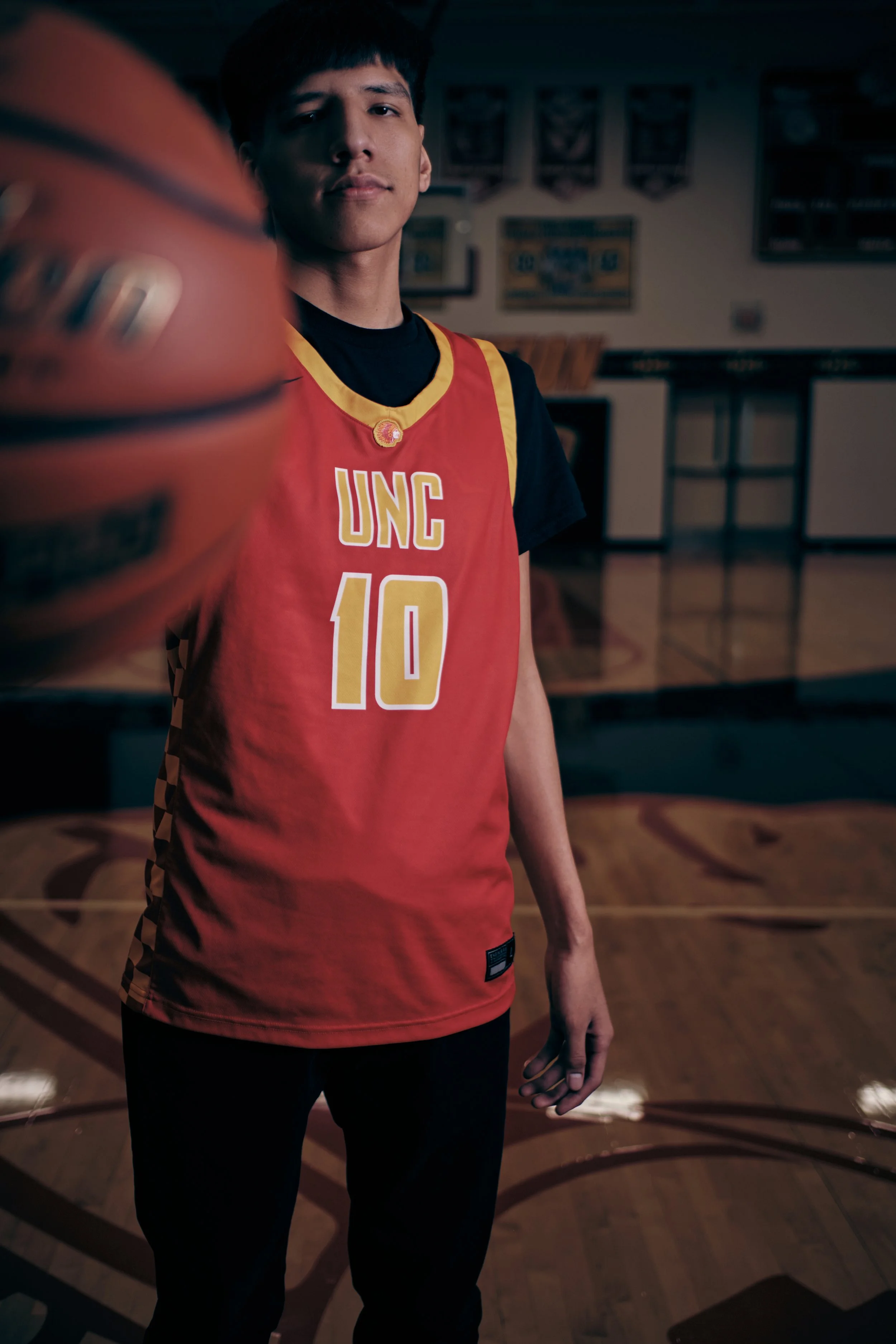 A young man in a red basketball jersey with 'UNC' and number 10 stands on a basketball court, holding a basketball in front of the camera with a blurred background of a gymnasium.