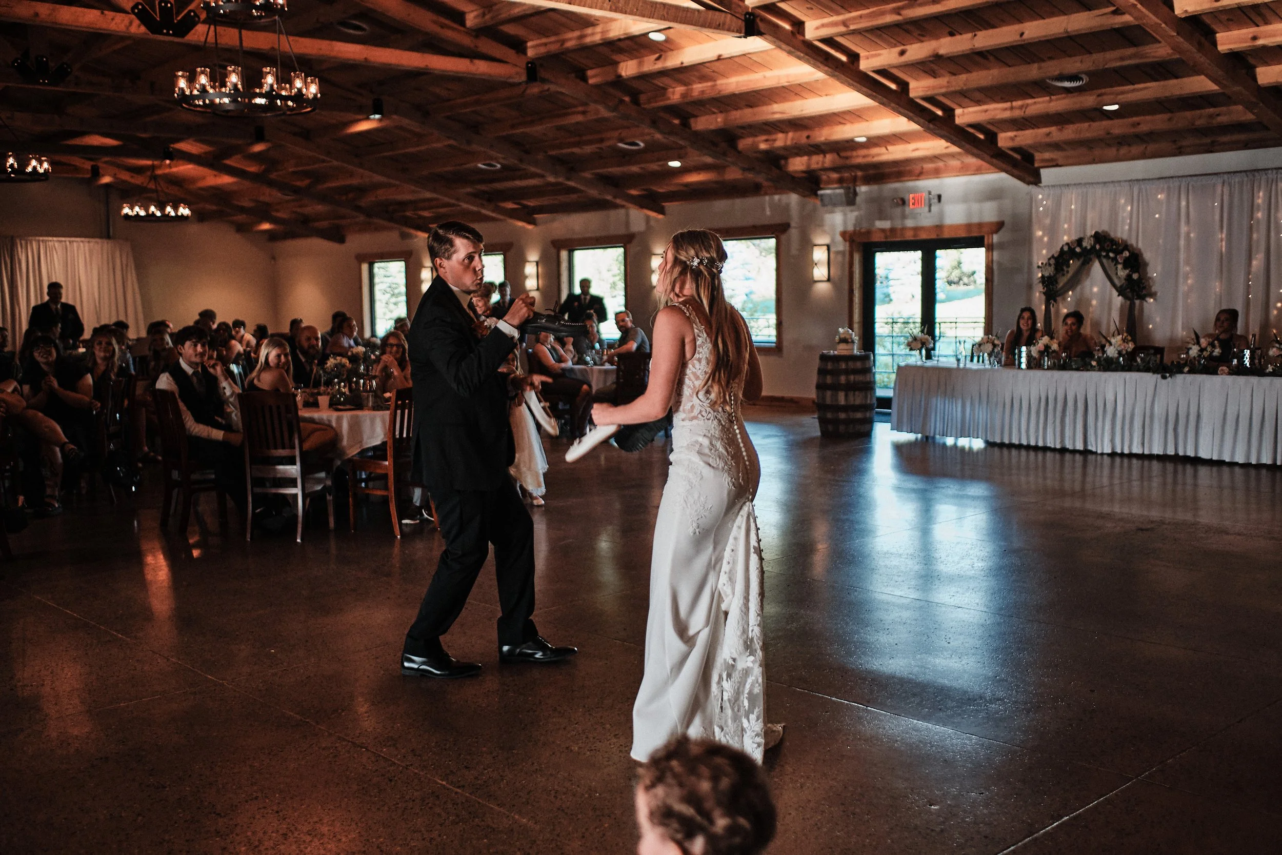 A wedding reception with a bride and groom dancing, surrounded by guests seated at tables, in a rustic venue with wooden ceiling and decorated with flowers and fairy lights.