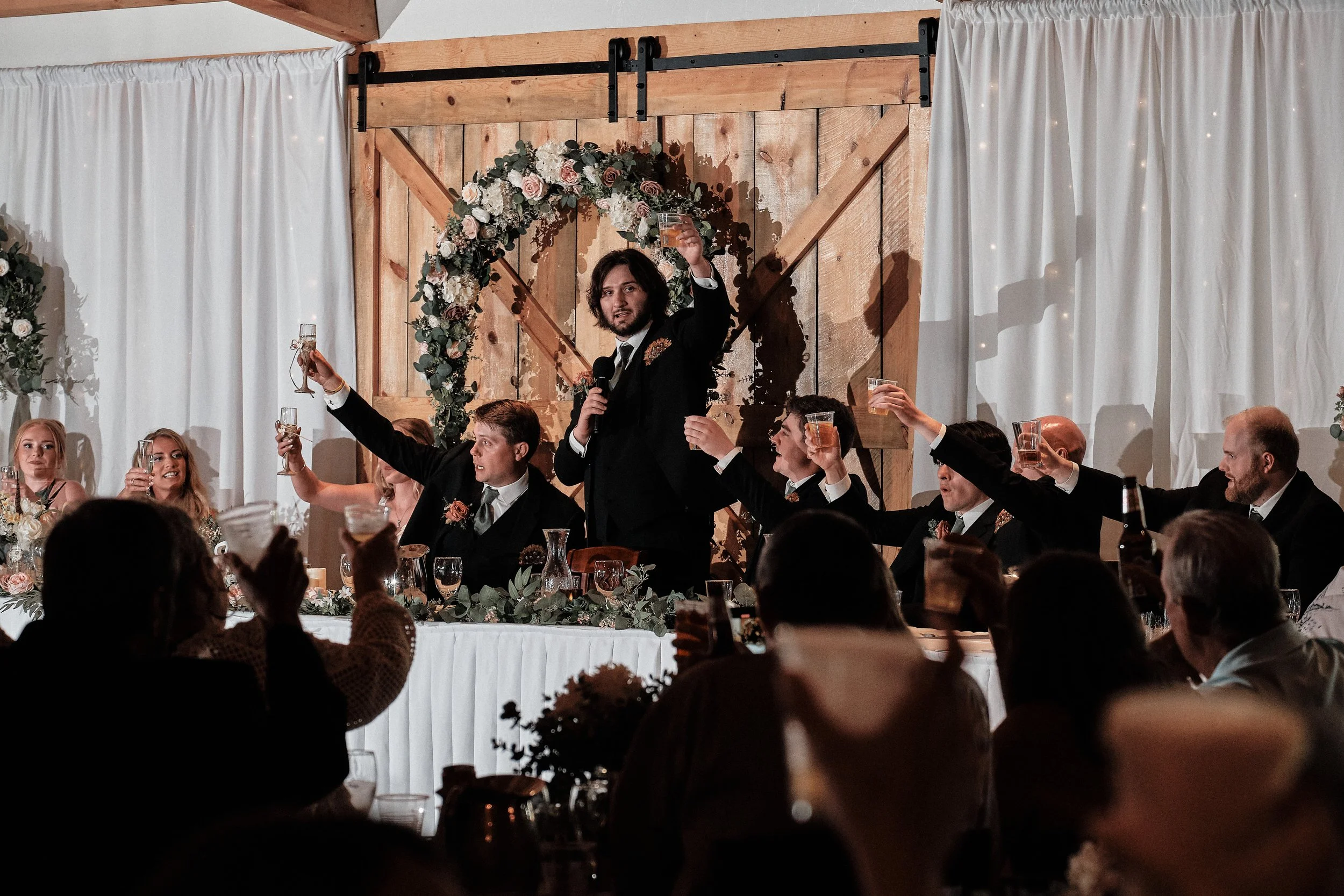 A man giving a toast at a wedding reception, standing behind a decorated table with guests raising glasses. The background features a wooden wall with a floral heart-shaped arch.
