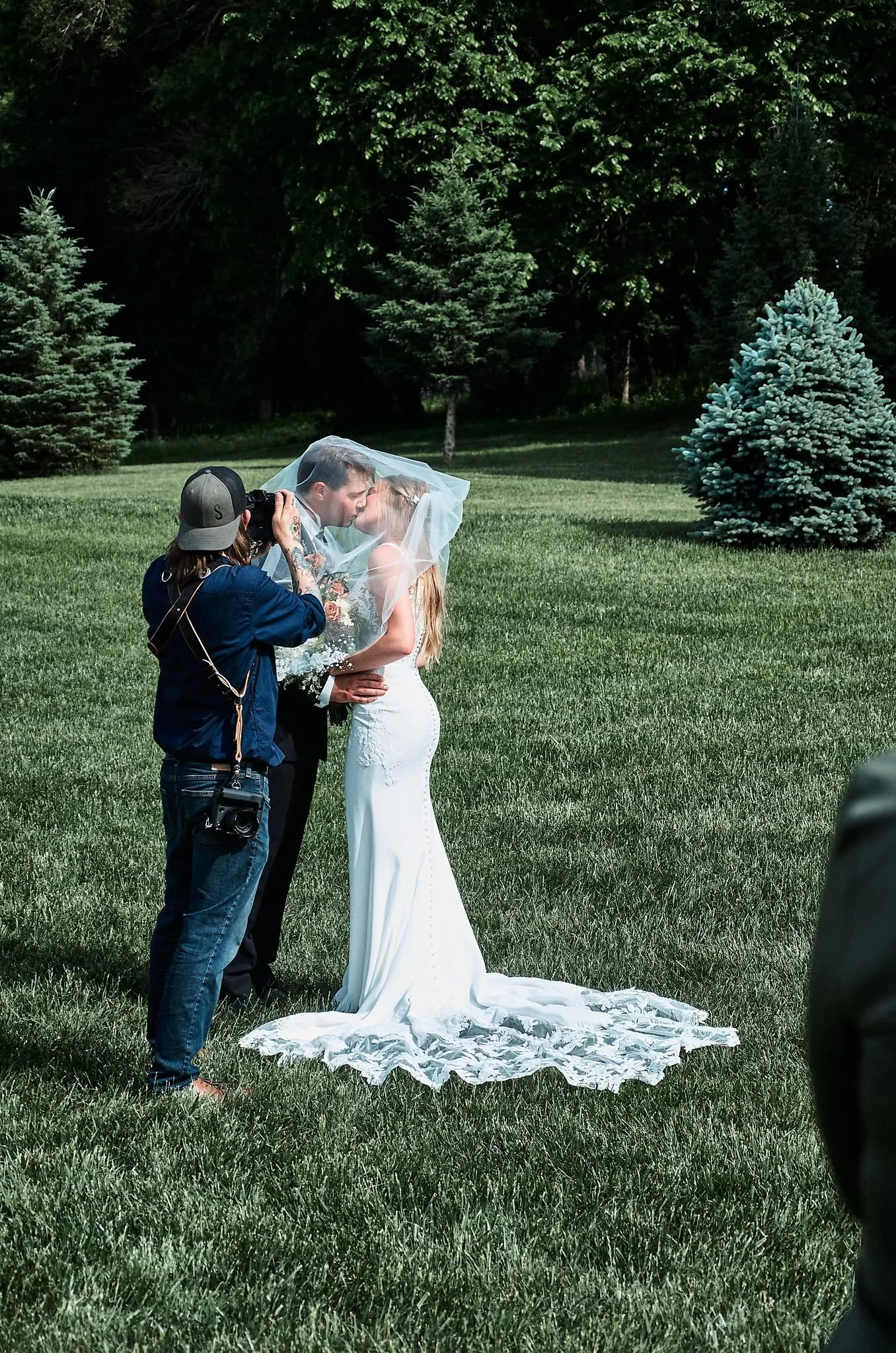 A bride and groom sharing a kiss under a veil outdoors on a grassy area with trees in the background. A photographer is taking their picture.