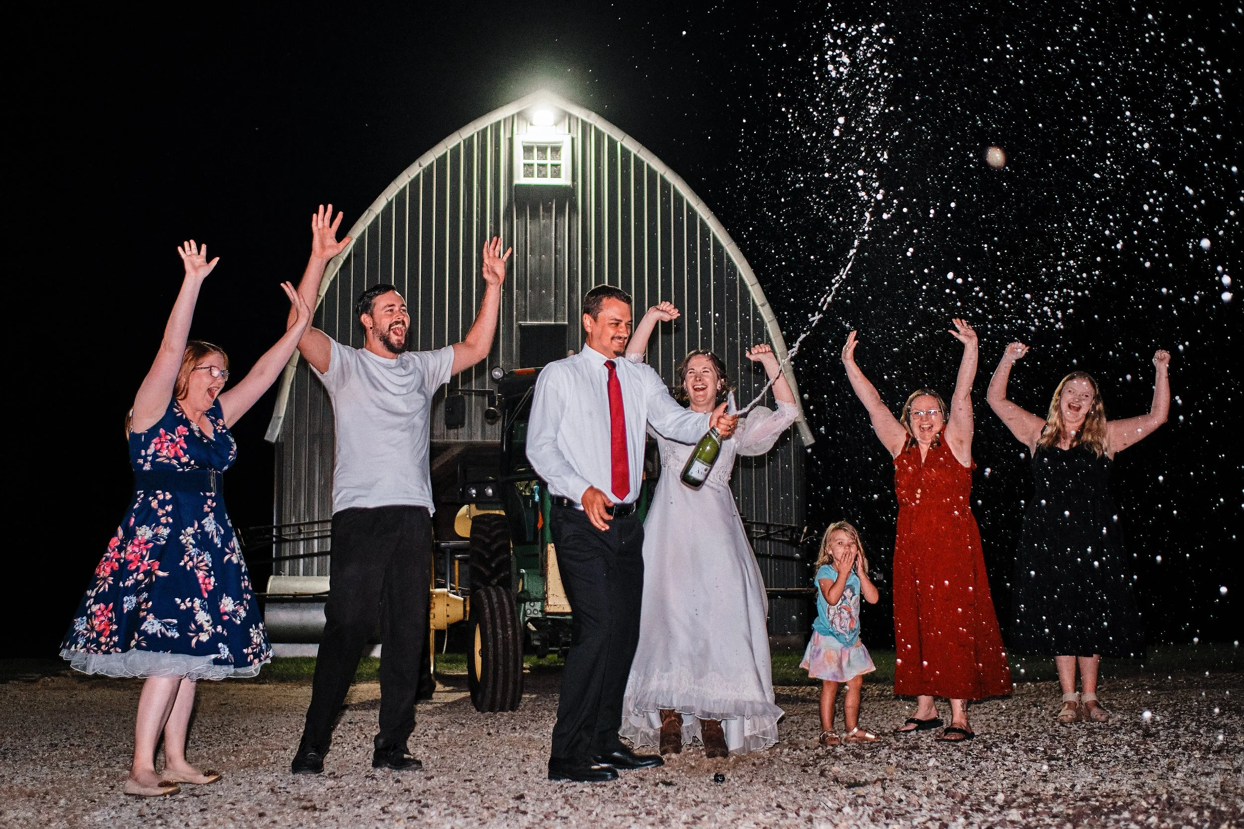 Group of people at night celebrating with champagne outside, some wearing fancy dresses, with a barn in the background, and snow falling.