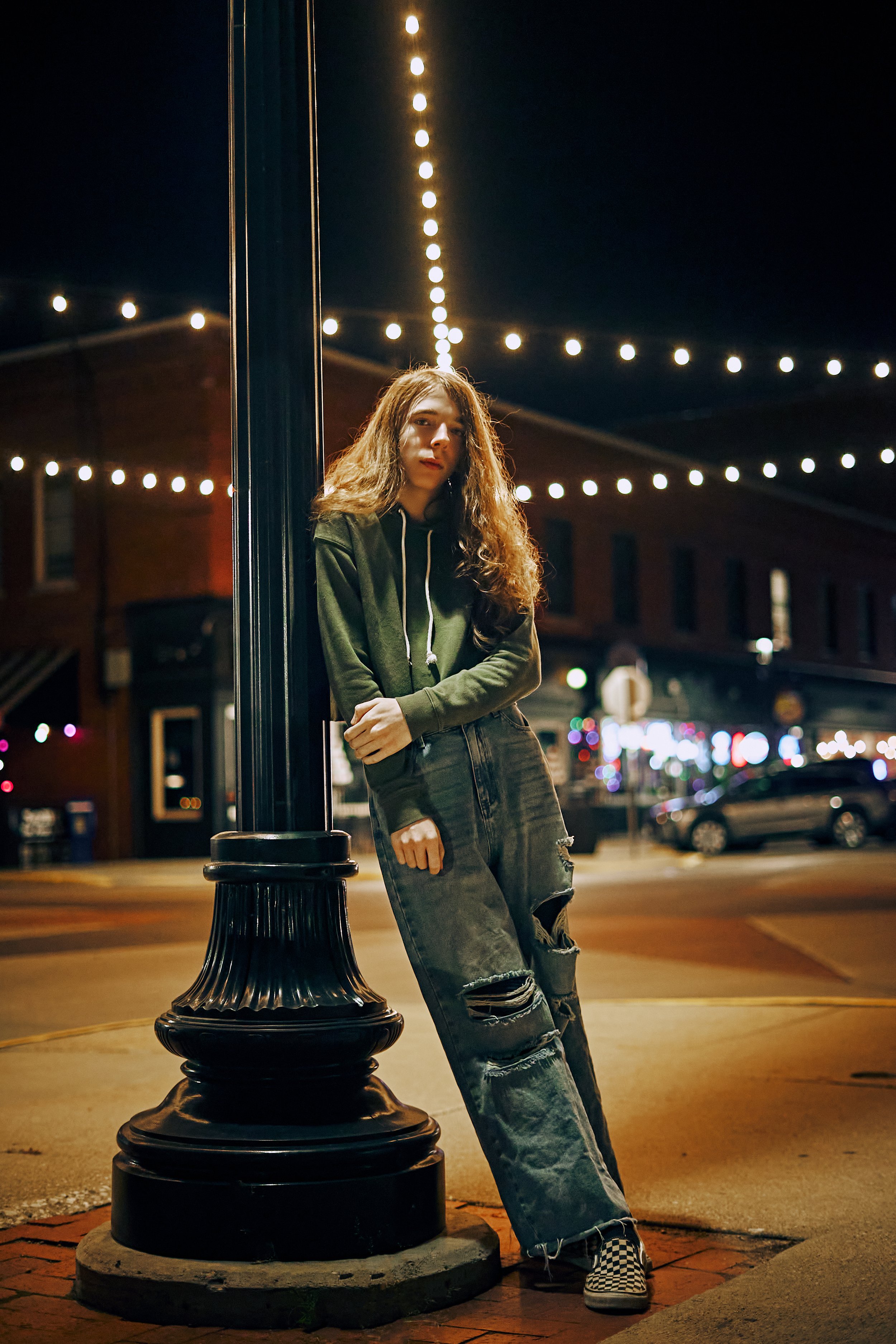 A young woman with long curly hair stands at night on a city street, leaning against a streetlamp with her arms crossed. She wears a green hoodie, distressed jeans, and checkered slip-on shoes. Strings of lights are overhead, and brick buildings with