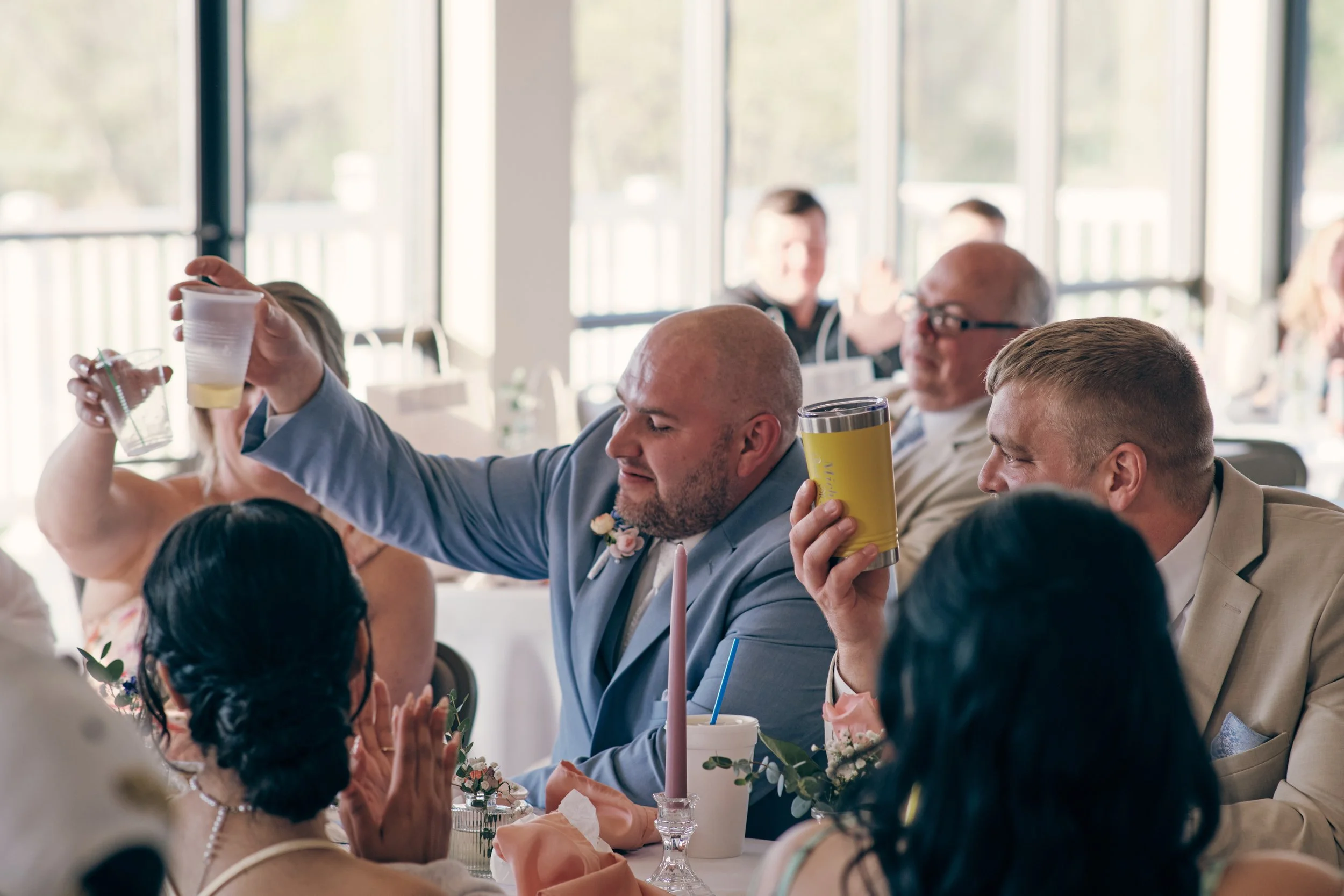 People at a wedding reception raising glasses in a toast indoors near large windows.