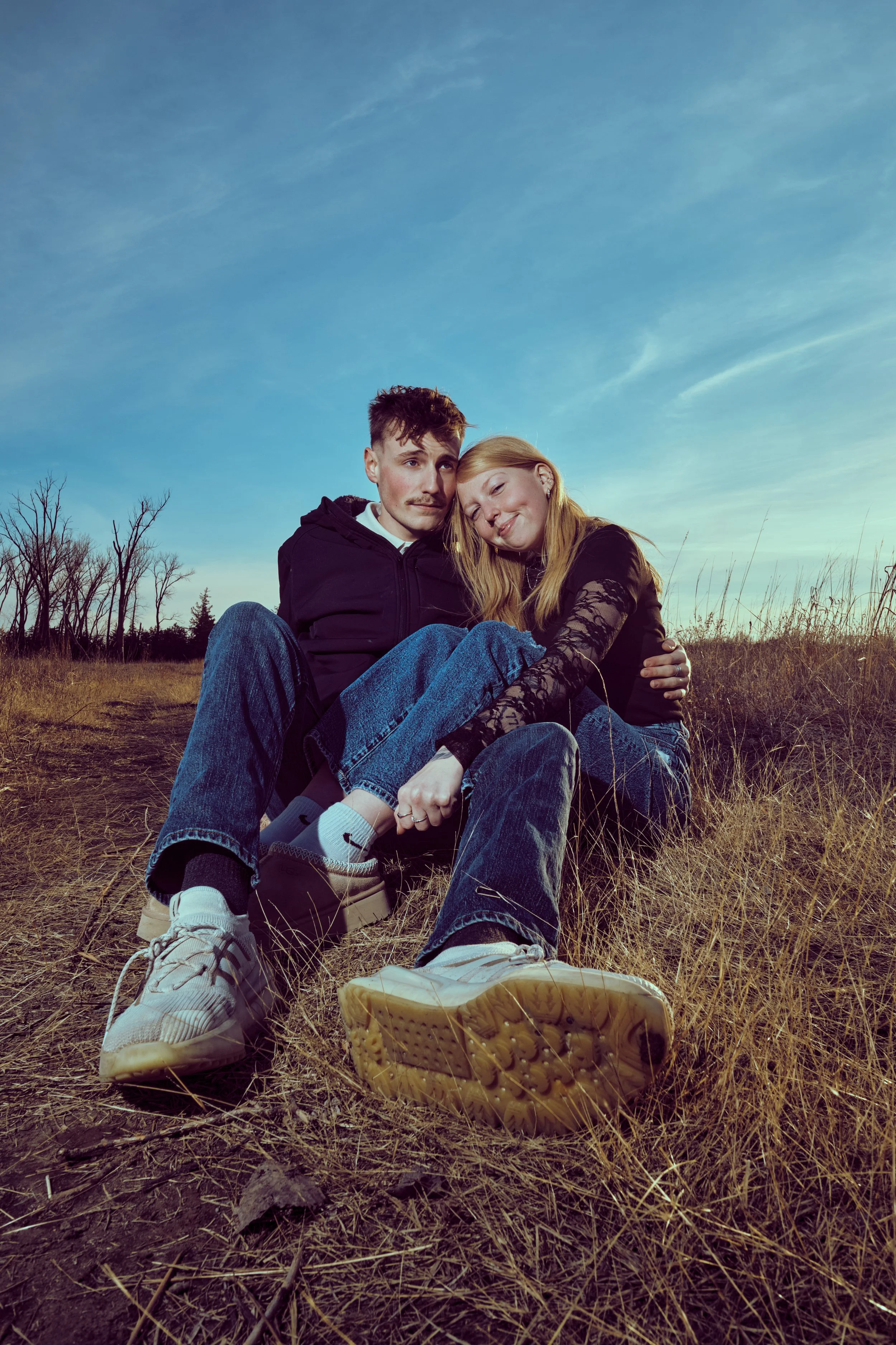 A young couple sitting together in a grassy field, smiling and embracing each other under a blue sky with some clouds. The person on the left has short brown hair, and the person on the right has long blond hair and is wearing a black top with lace s