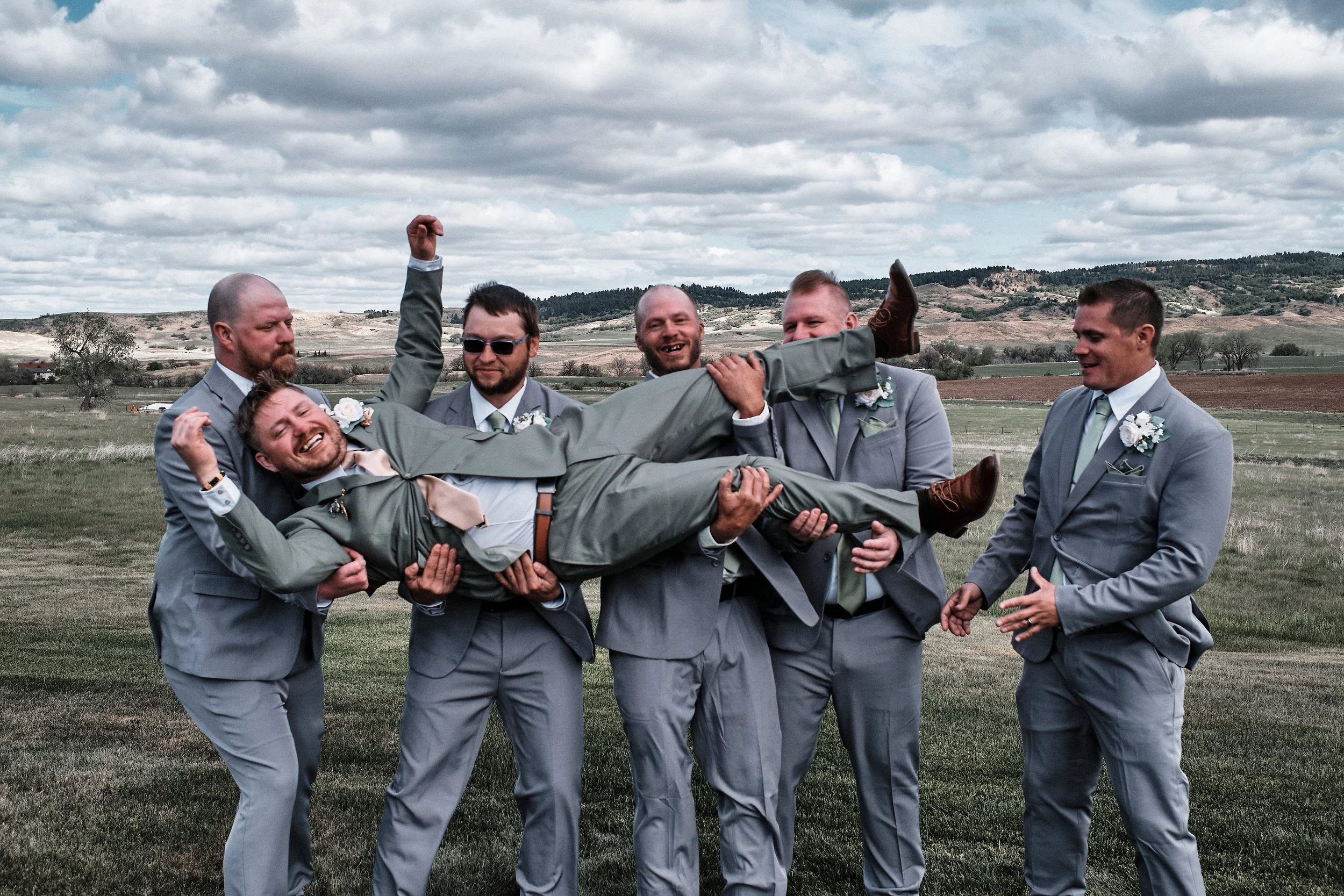 A group of men in suits celebrating outdoors, with one man being lifted horizontally by the others, in a rural landscape under a cloudy sky.